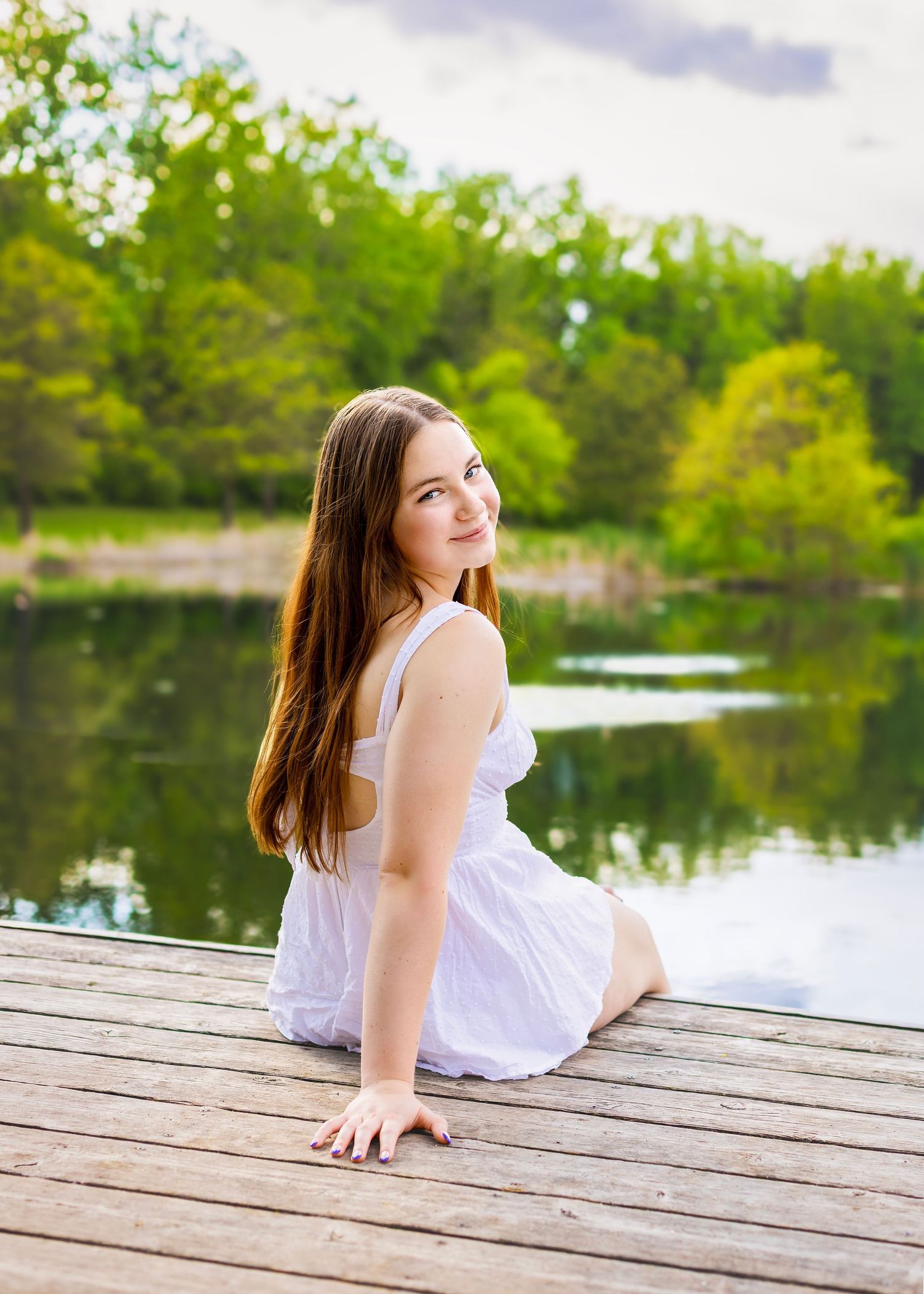 Young woman in white dress sitting on wooden dock by serene lake surrounded by lush green trees.