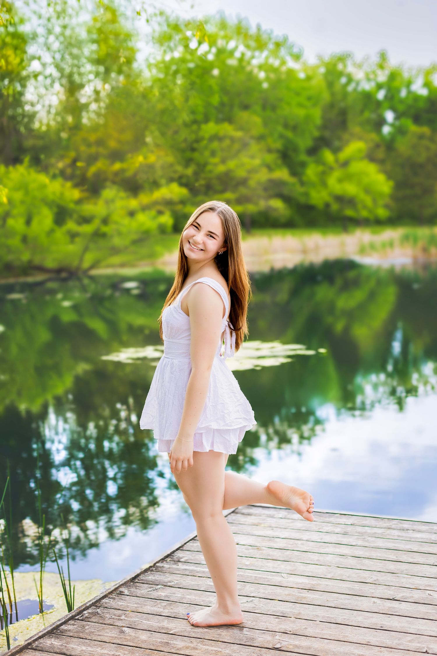 Young woman in white dress standing barefoot on wooden dock by a serene green lake on a sunny day.