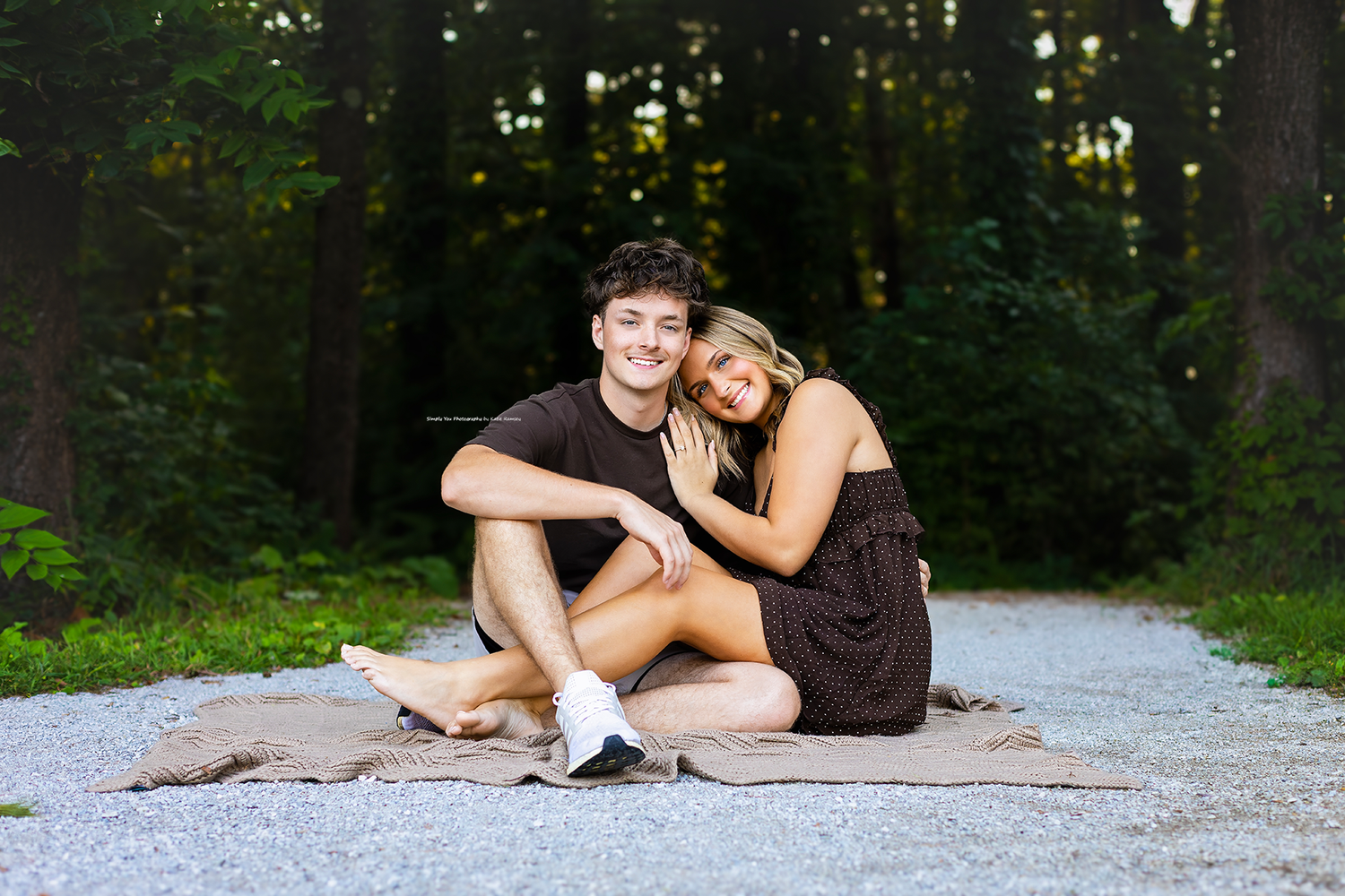 A smiling couple sits together on a blanket on a gravel path surrounded by lush green trees.
