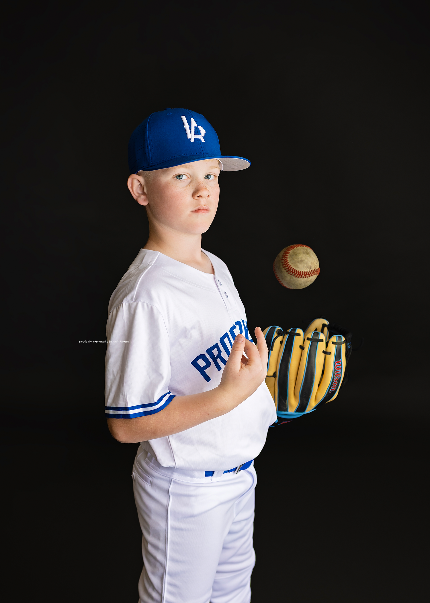 Young baseball player in white Prospects uniform and blue LA cap tossing a ball with yellow glove on black background.