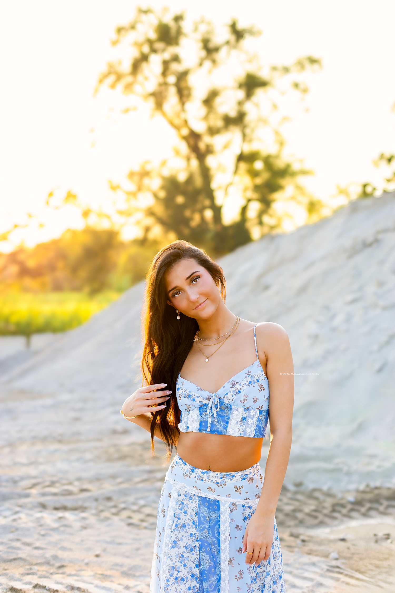 Young woman in blue floral crop top and wide-leg pants poses outdoors at golden hour with trees in background.