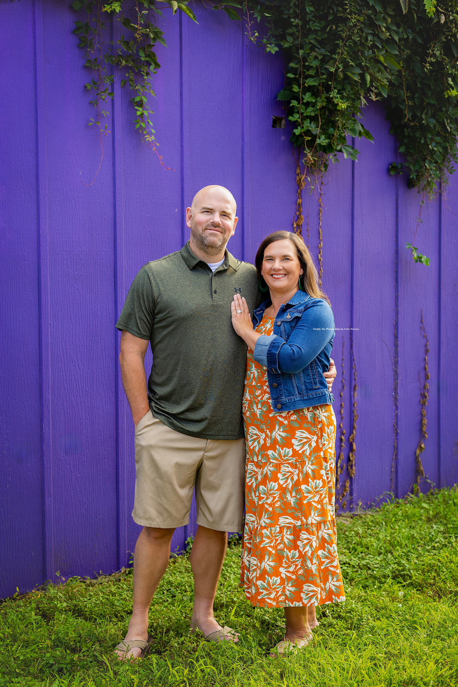 Smiling couple posing in front of a vibrant purple fence with ivy, man in green polo and woman in orange floral dress.