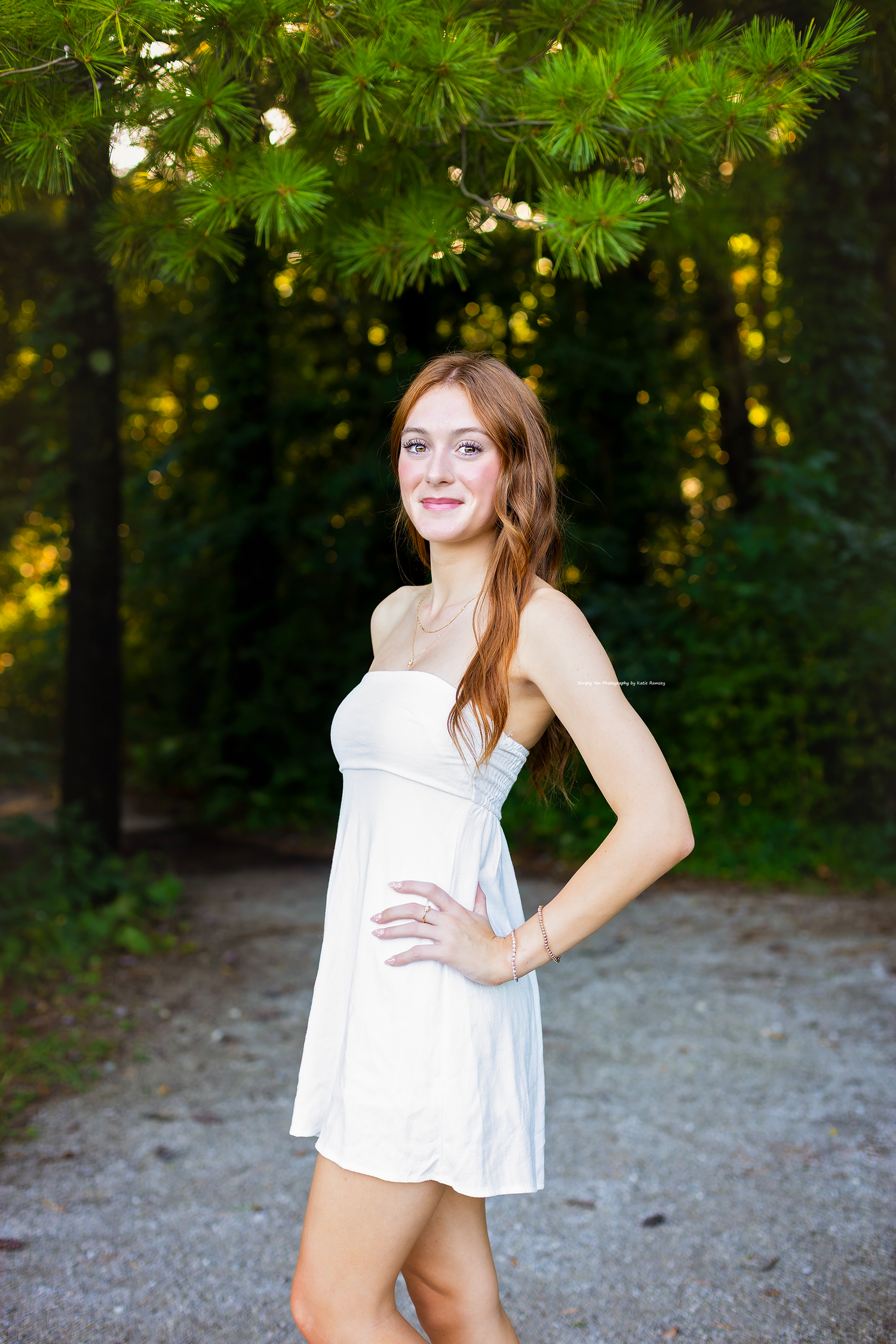 Young woman in white strapless mini dress posing on a forest path surrounded by green pine trees.