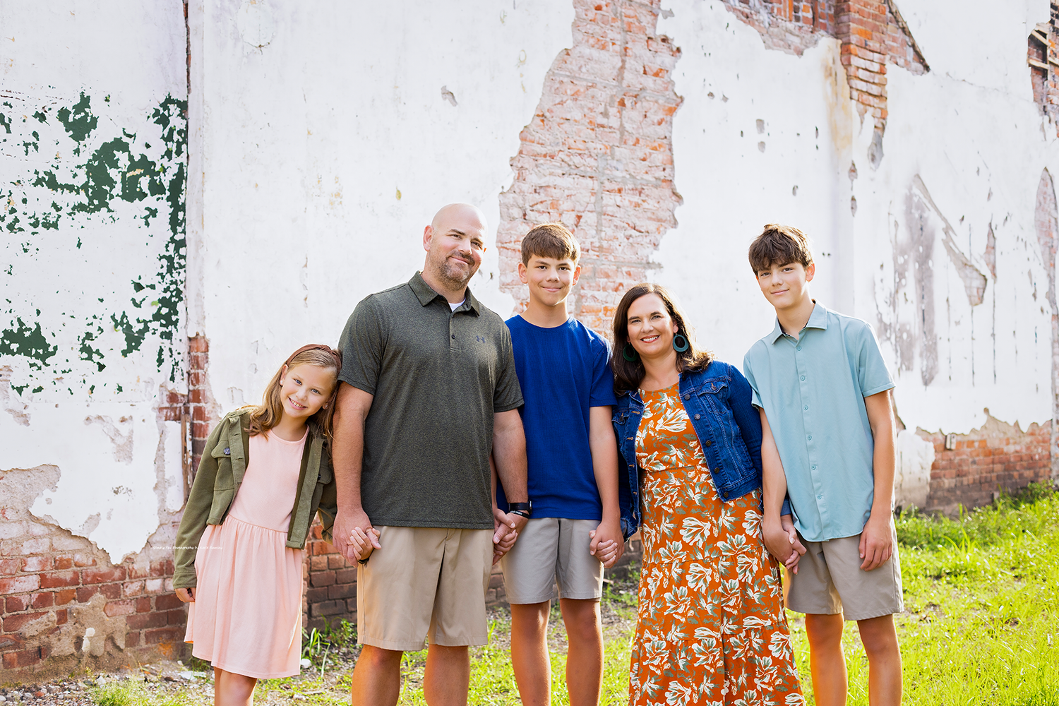 Family of five posing outdoors near a rustic brick wall, wearing colorful casual outfits, smiling together on a sunny day.