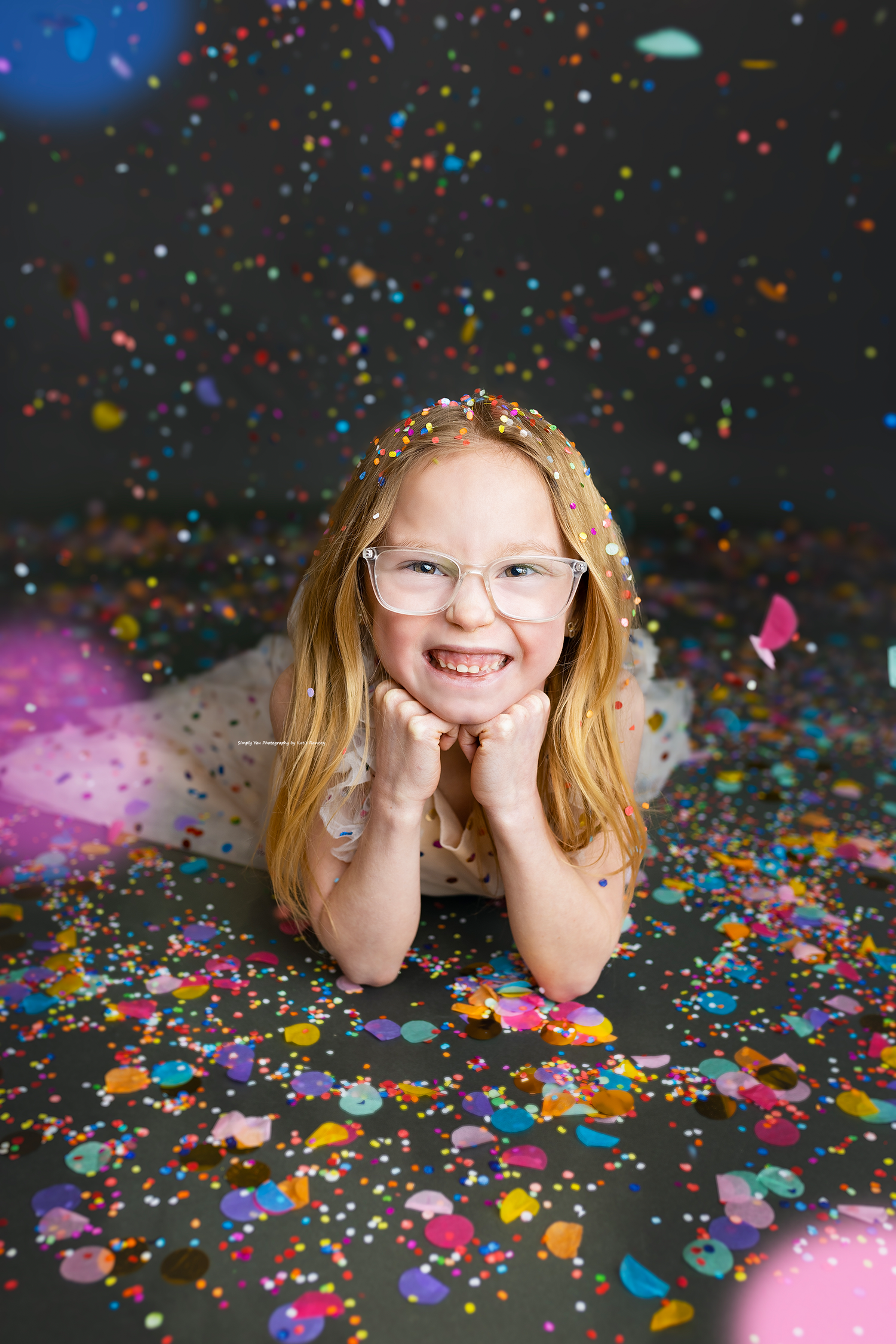 Adorable young girl with glasses lying on the floor smiling amid scattered colorful confetti in a studio.
