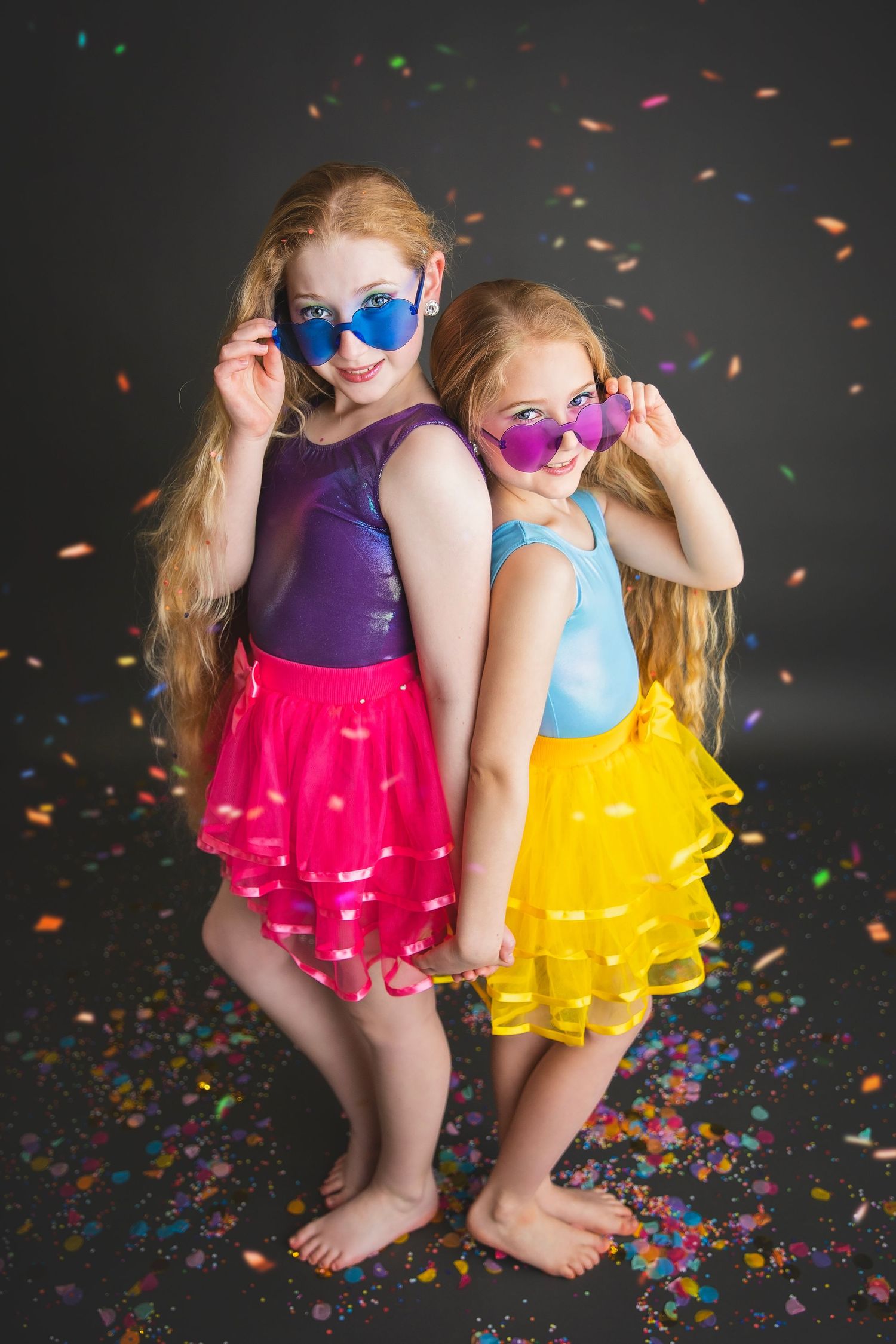 Two young girls in colorful tutus and heart-shaped sunglasses pose back-to-back with confetti falling around them.