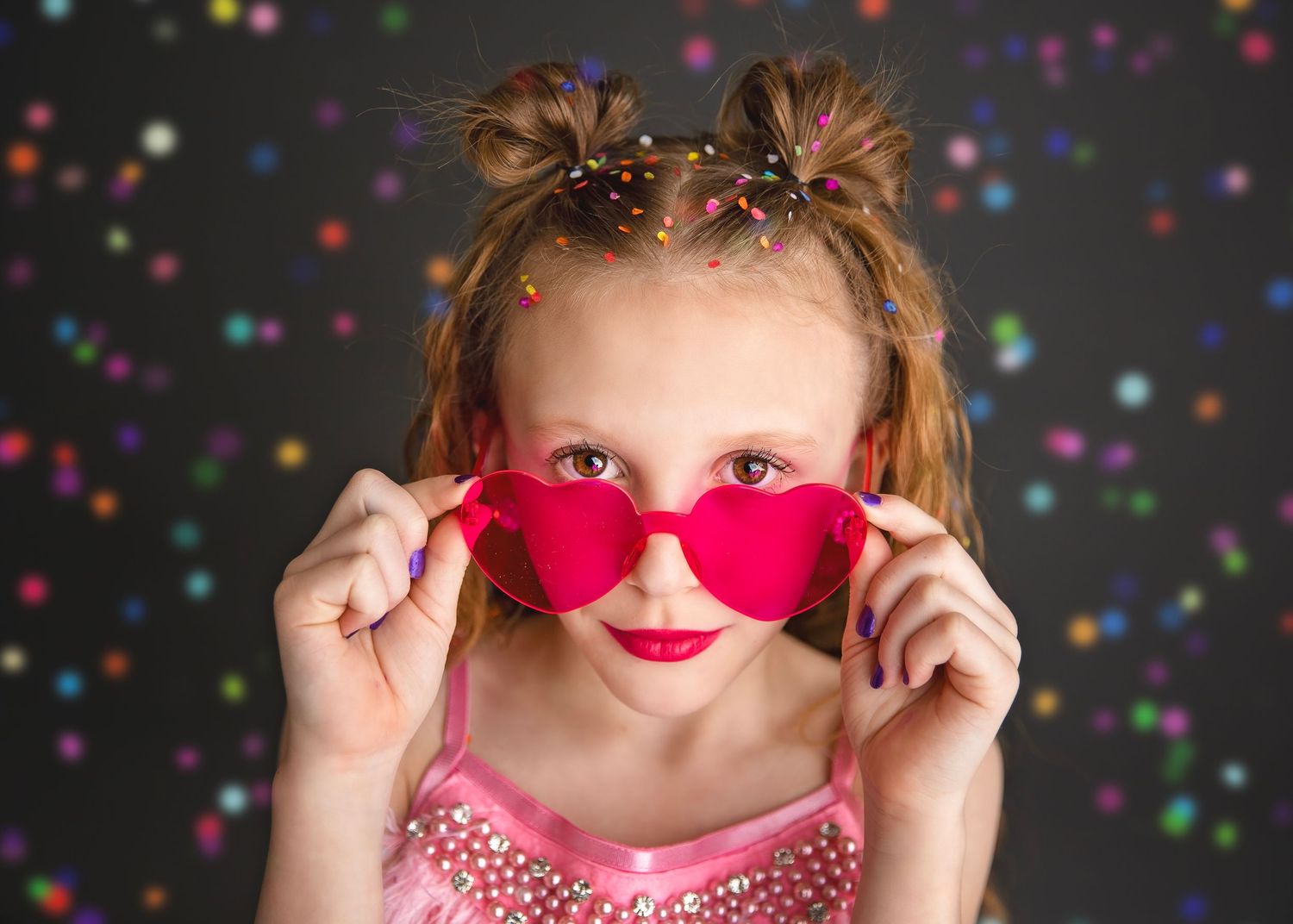 Girl in pink dress holding red heart-shaped sunglasses with confetti background.