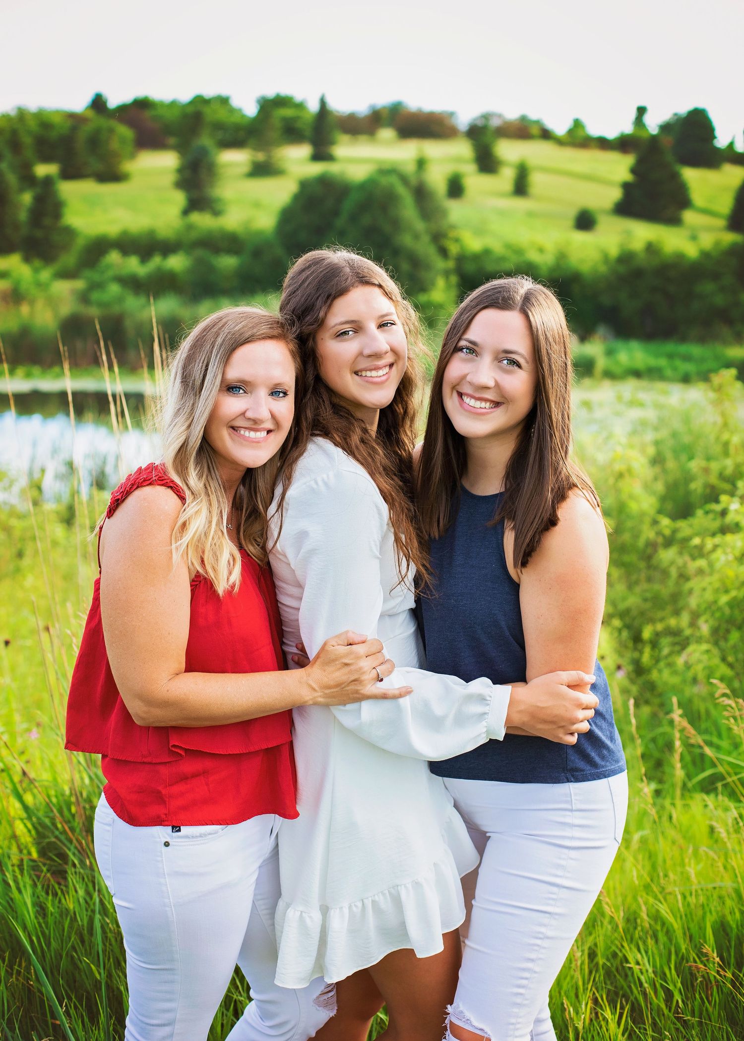 Three young women smiling and embracing outdoors with green hills and pond in background.