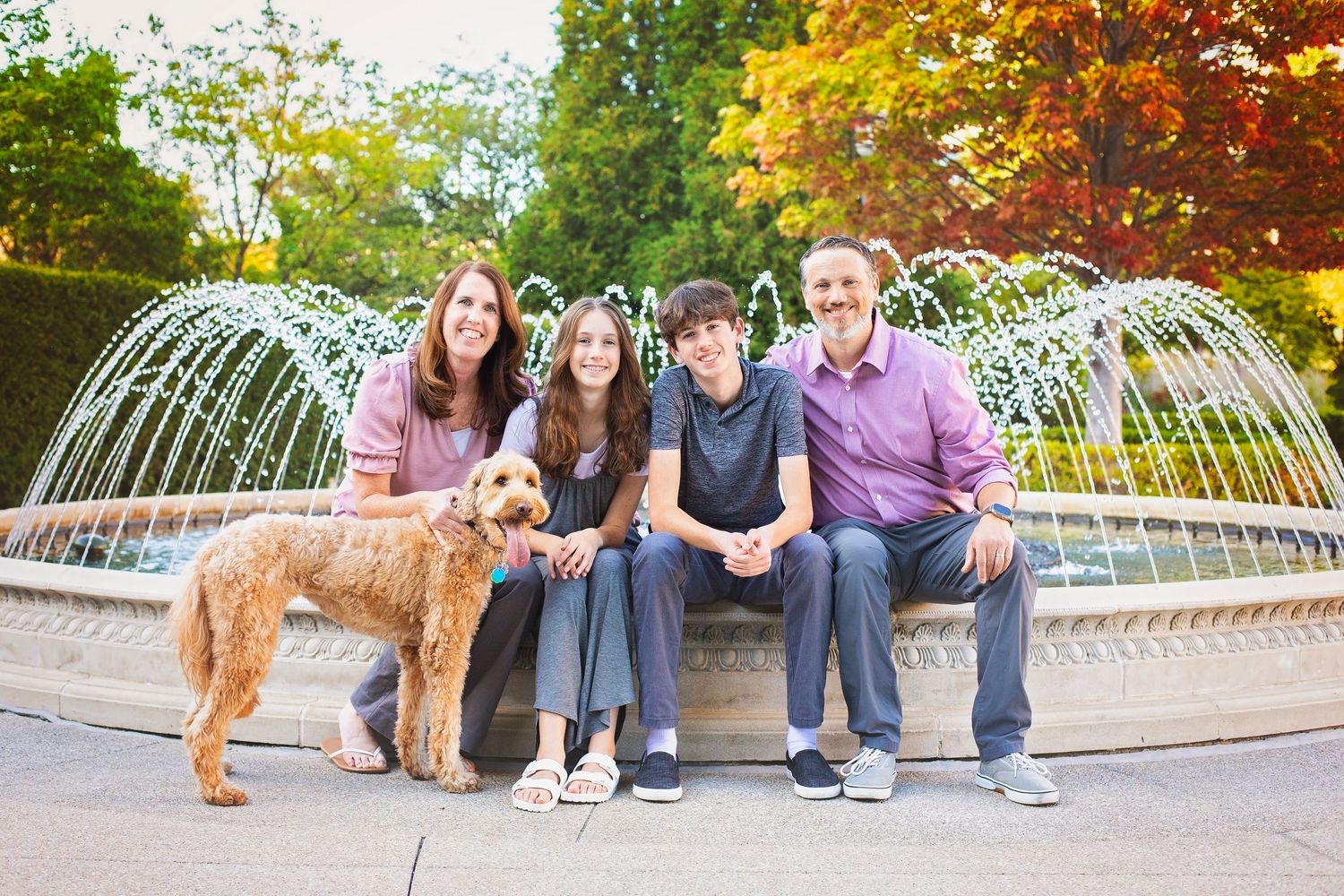 Family with a golden doodle sitting by a fountain in a park in autumn.