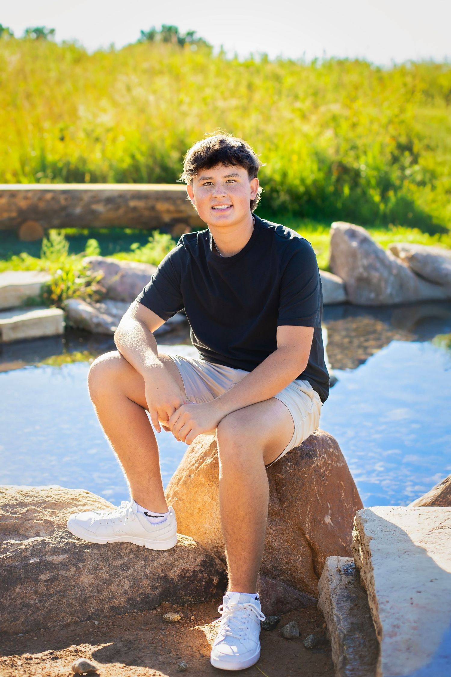 Smiling teenage boy sitting on rocks near a pond surrounded by lush green grass in sunlight.