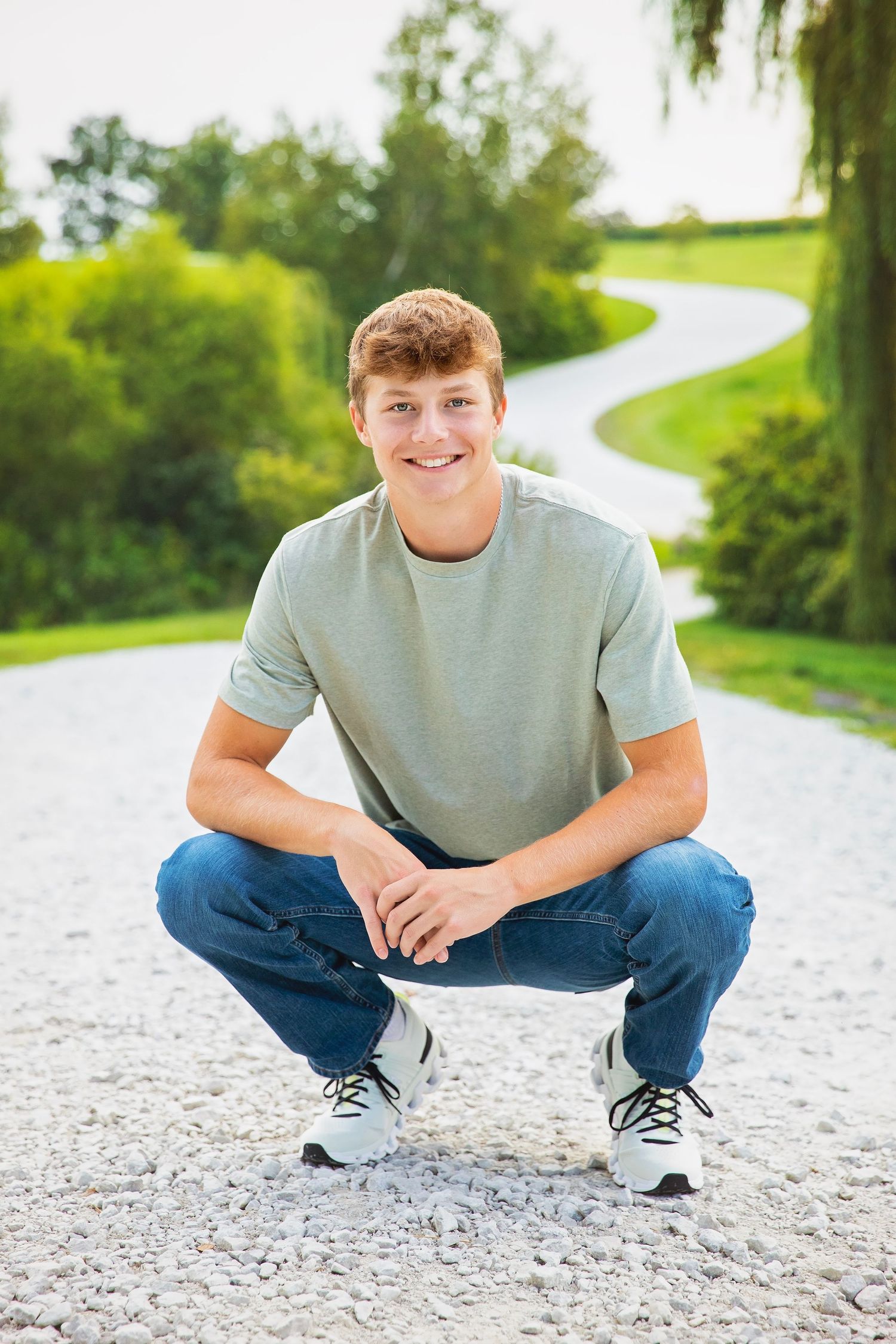 Smiling teenage boy crouching on a gravel path outdoors in a sage green t-shirt and jeans.
