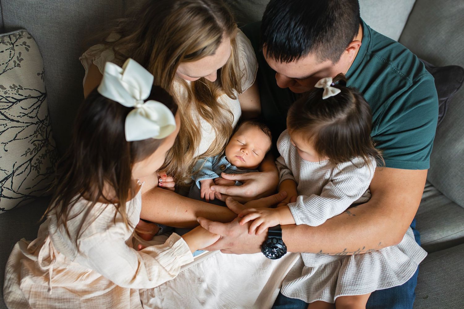 Family of five gathered on a gray sofa, parents and two daughters lovingly admiring their newborn.