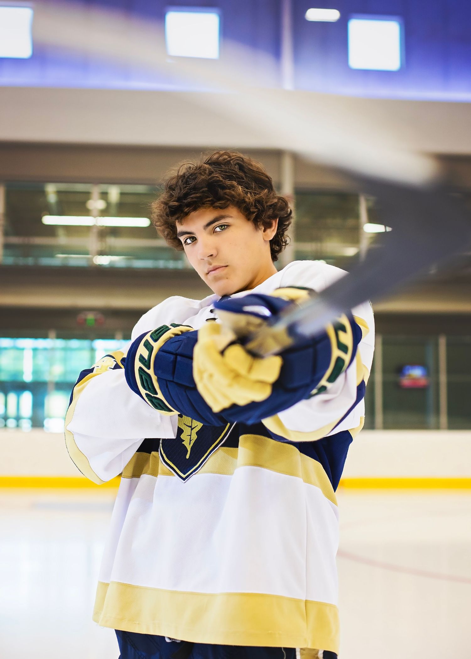 Teen hockey player in navy and gold jersey points stick toward camera on indoor ice rink.