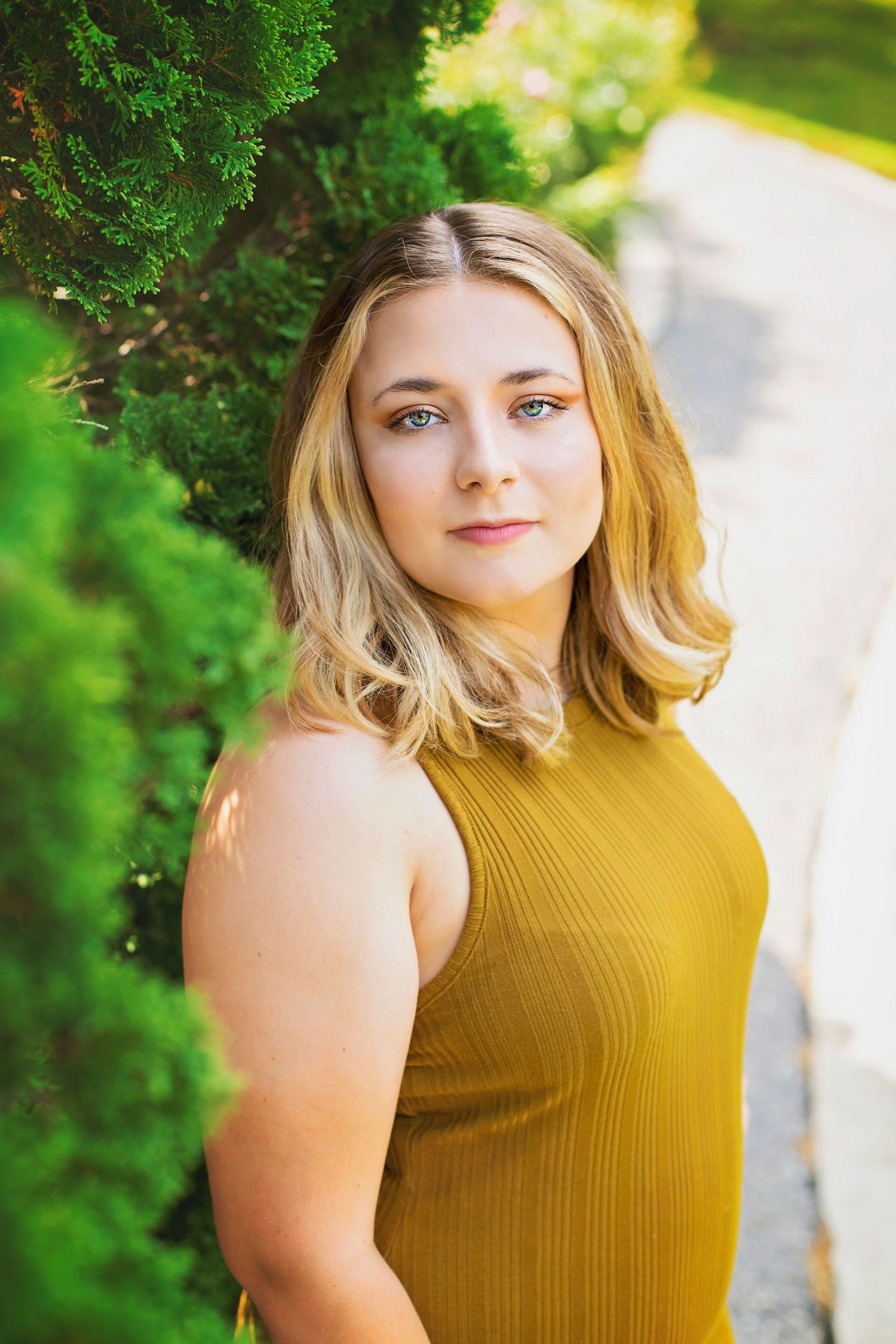 Young blonde woman in mustard yellow sleeveless top posing beside green shrubs outdoors.