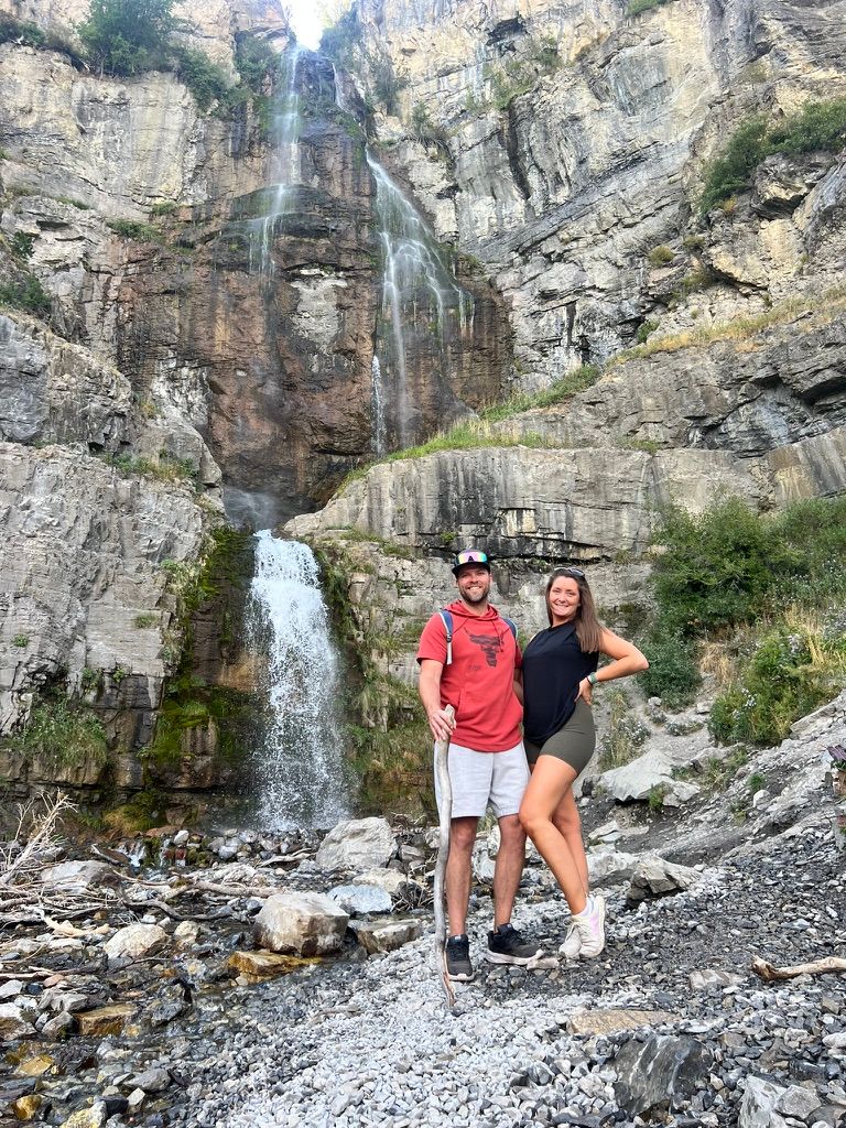 Couple posing in front of tall waterfall cascading down rocky canyon cliff face.