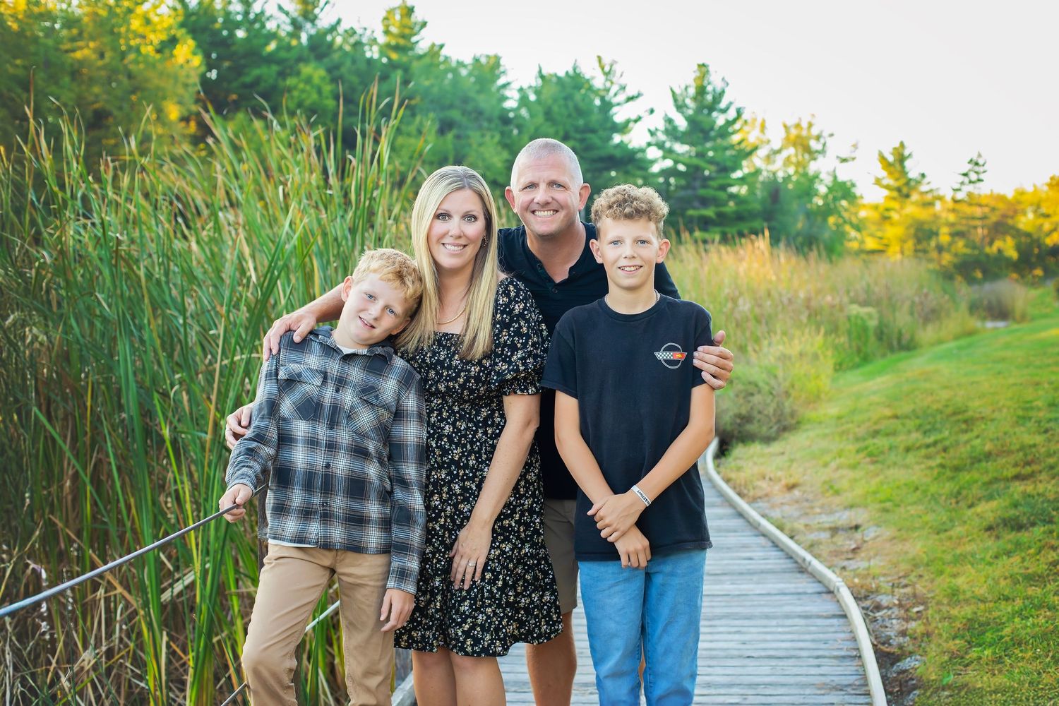 A smiling family of four poses together on a wooden boardwalk path surrounded by tall golden grasses.