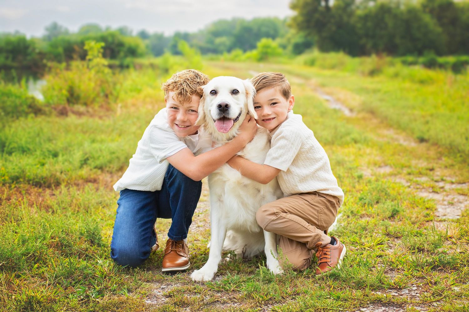 Two young boys hugging a happy golden retriever dog in a lush green outdoor field.