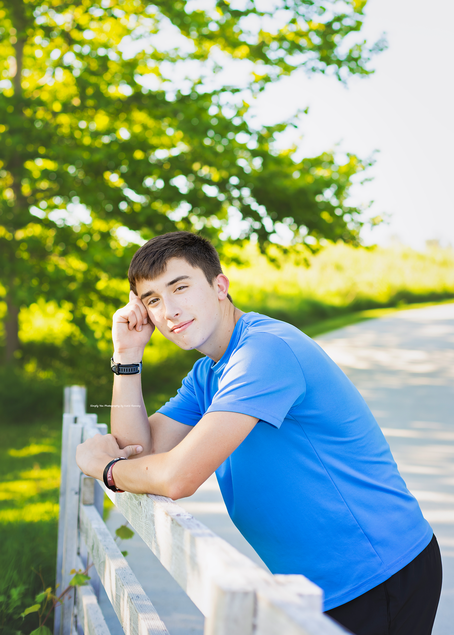 Teen boy in blue shirt leaning on white fence in sunny park setting.