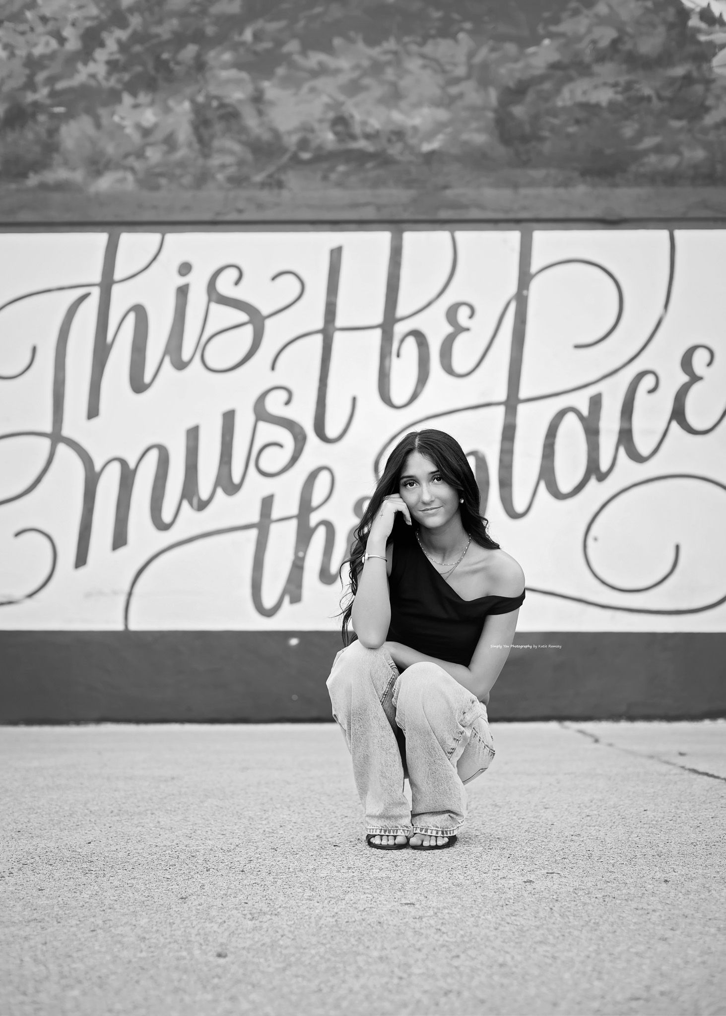 Young woman crouching in front of a mural reading This Must Be The Place, black and white photo.