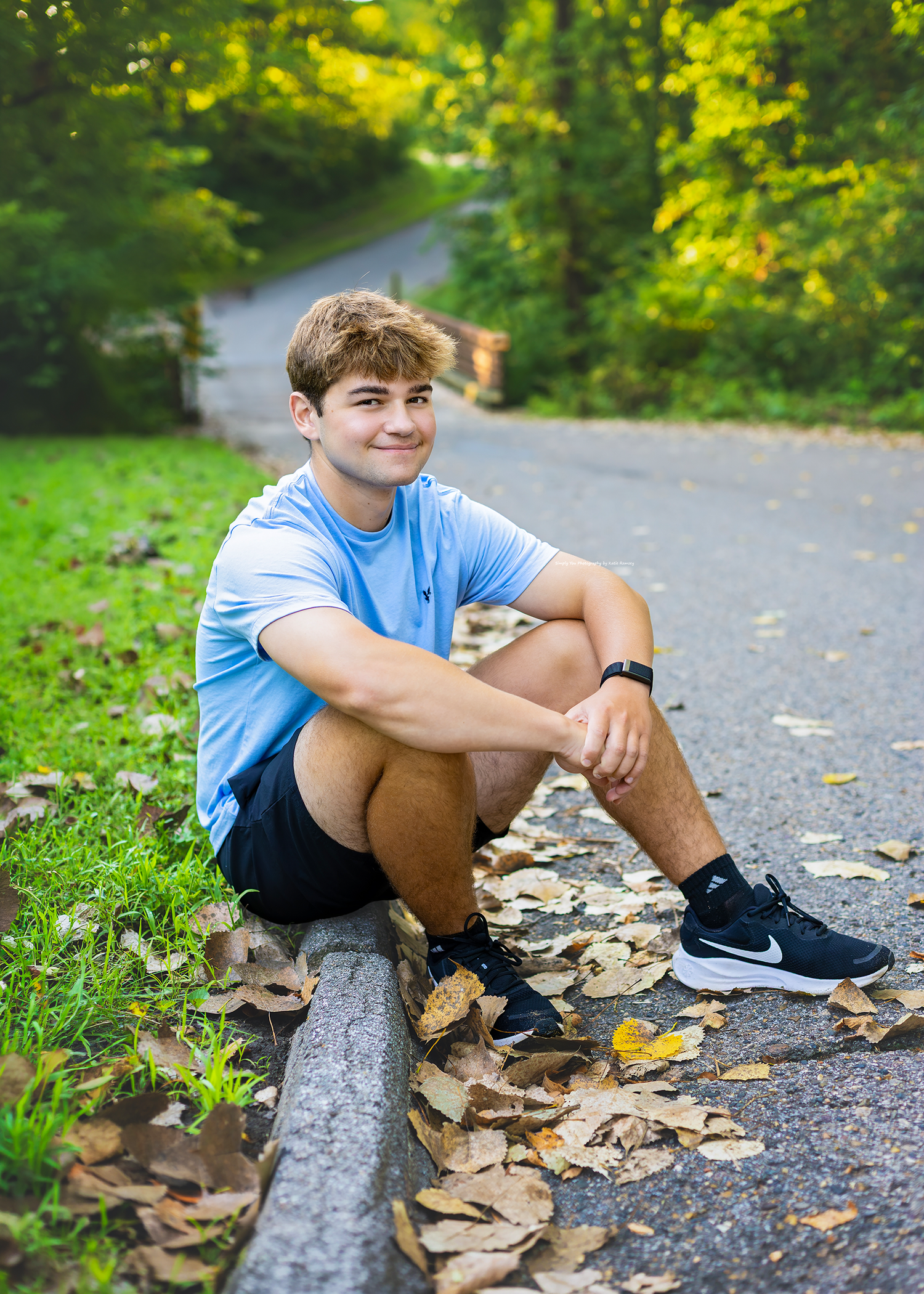 A smiling teenage boy sits on a curb surrounded by fallen leaves, wearing a light blue shirt, black shorts, and Nike sneakers.