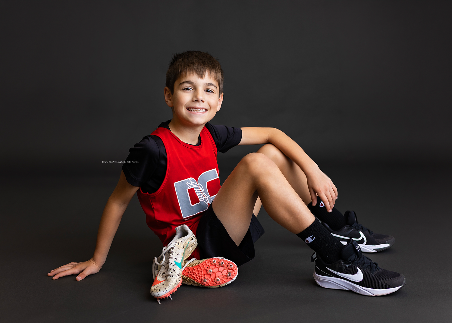 Smiling boy in red DC tank top sitting with track spikes, studio photo.