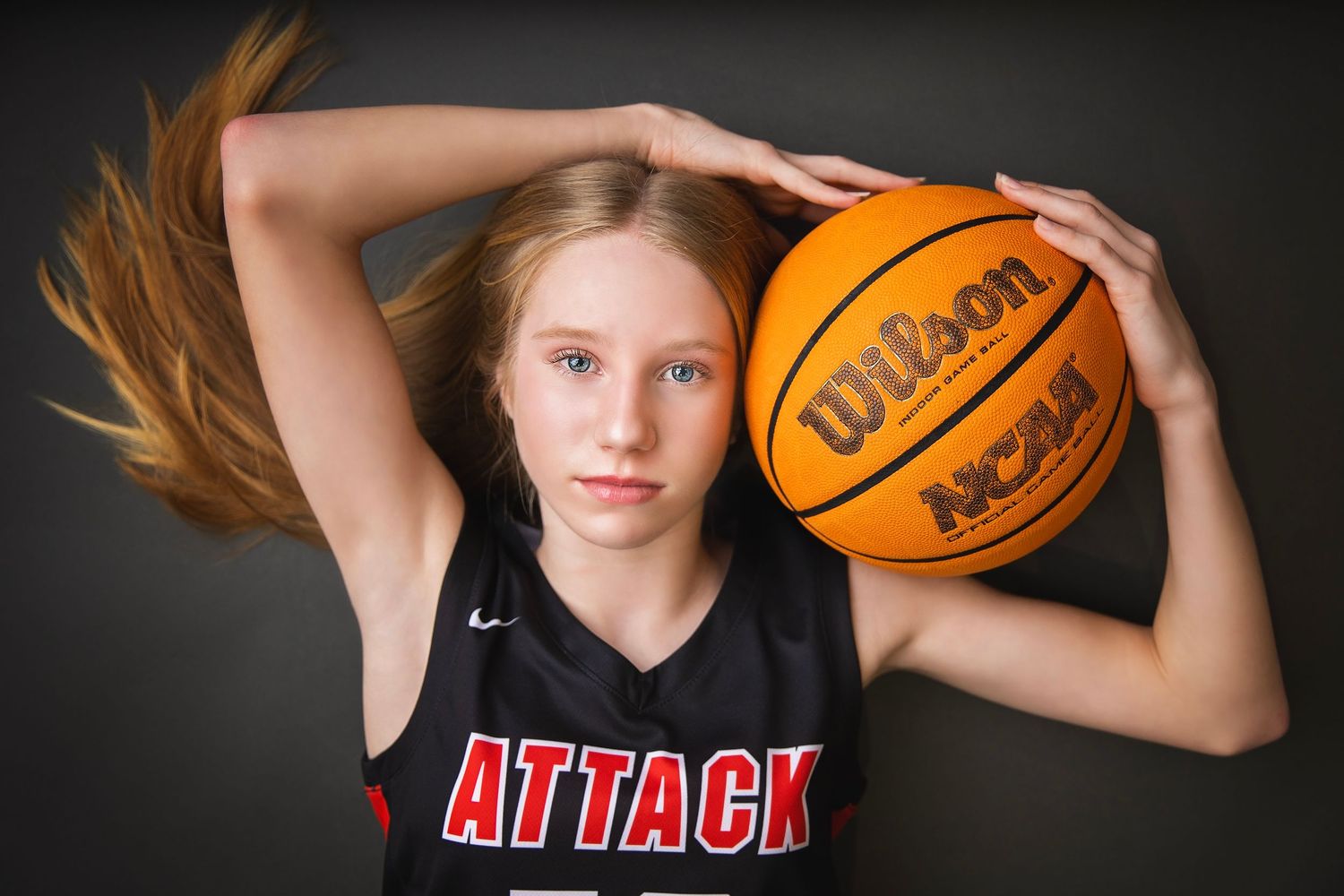 Blonde girl in Attack jersey balancing Wilson NCAA basketball overhead.