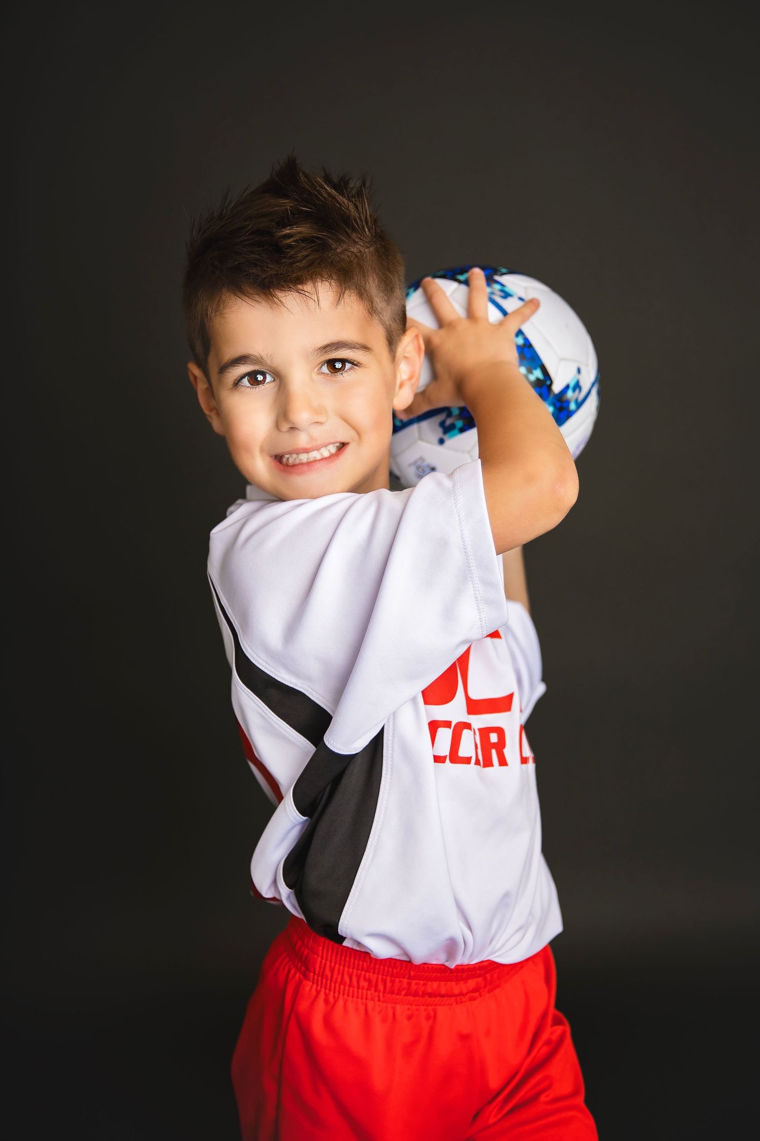 Smiling young boy in DCG Soccer Club uniform holds soccer ball overhead in dynamic sports portrait.