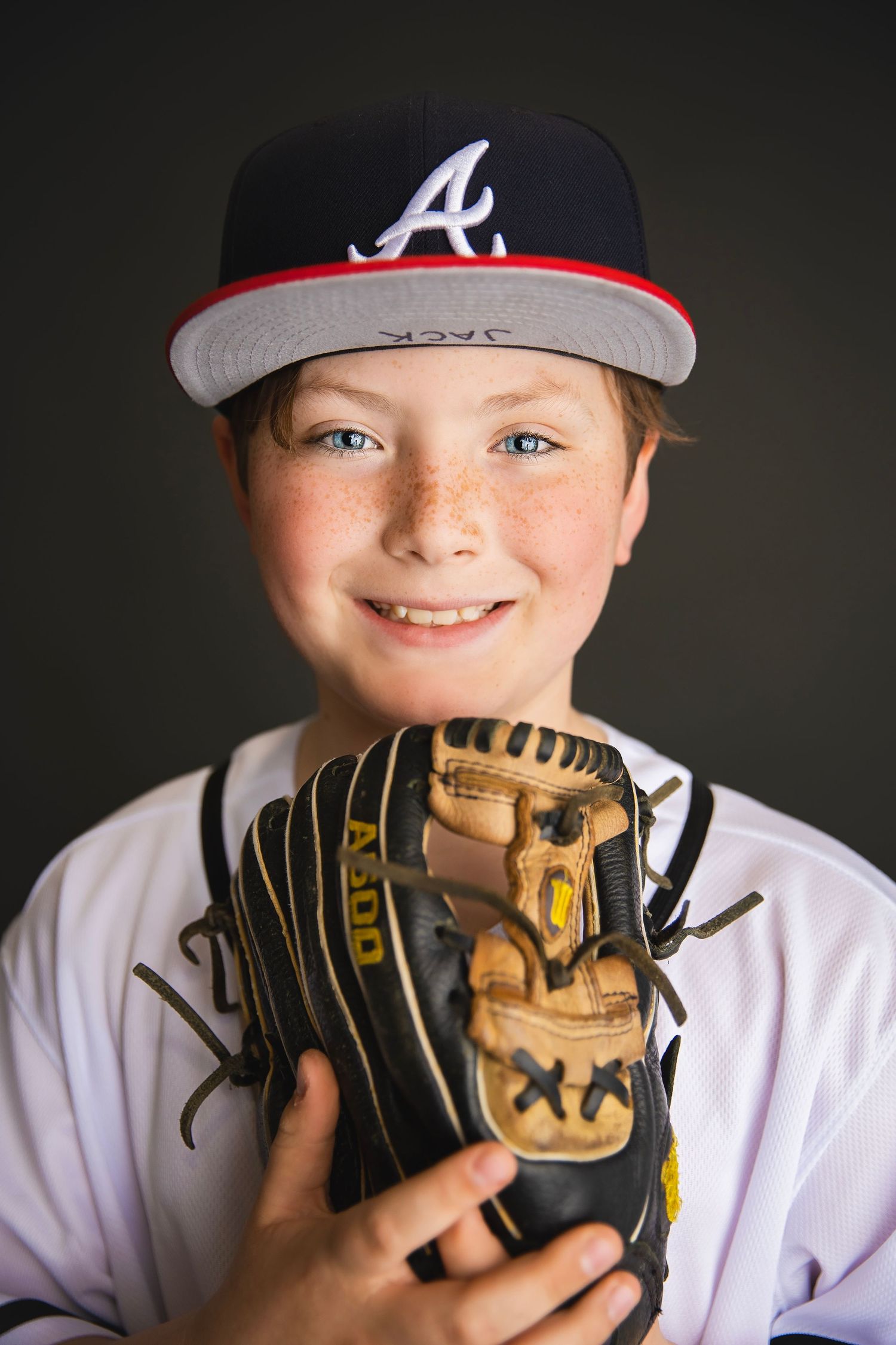 Smiling redhead boy in Atlanta Braves cap holding baseball glove toward camera, freckles, dark background.