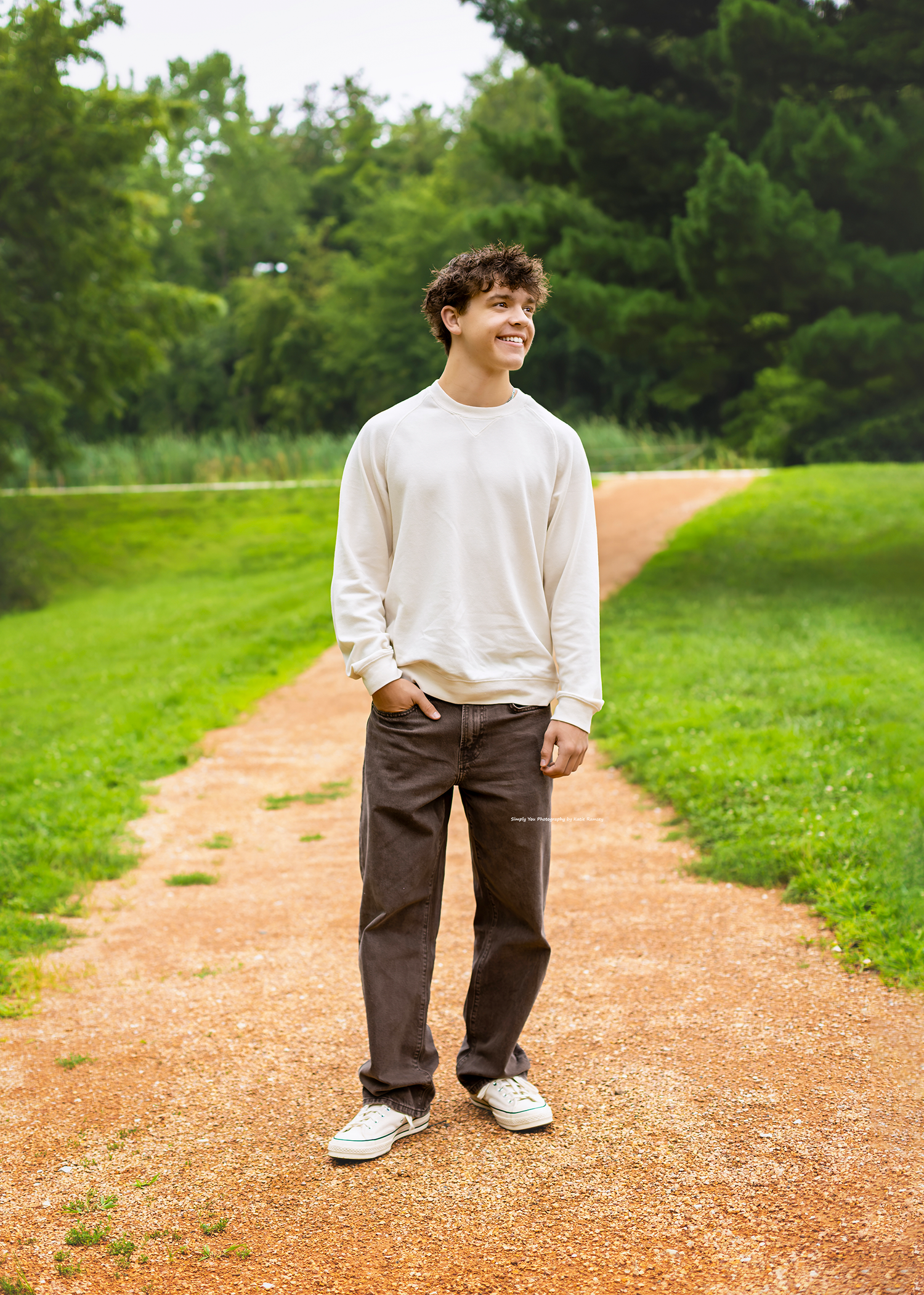 Young man in white sweatshirt and dark pants smiling on a dirt path surrounded by lush green grass and trees.