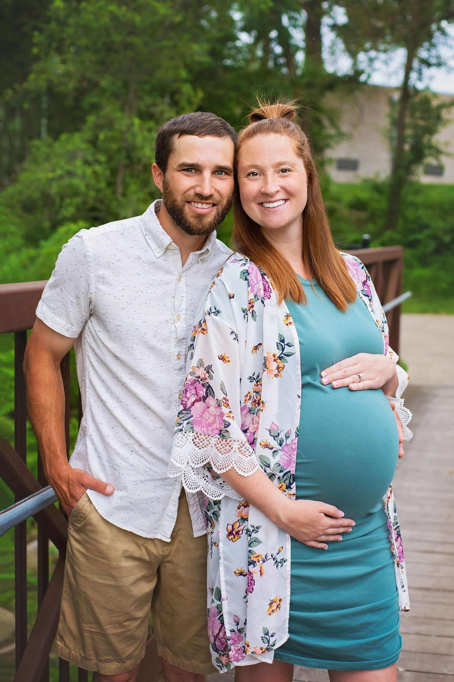 Smiling couple poses outdoors on a bridge, with pregnant woman in teal dress and floral kimono cradling her baby bump.