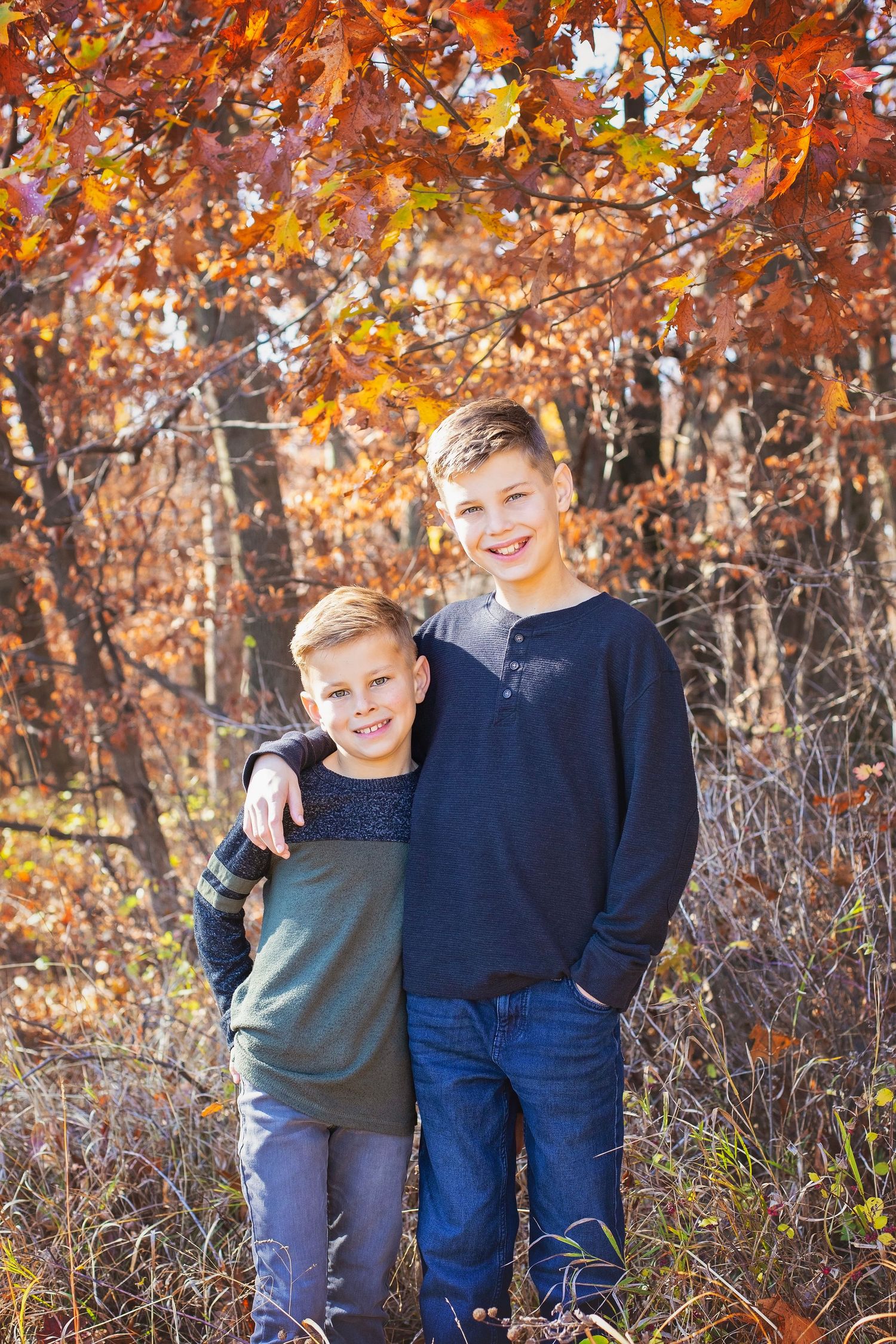 Two smiling brothers posing under vibrant red fall leaves outdoors.