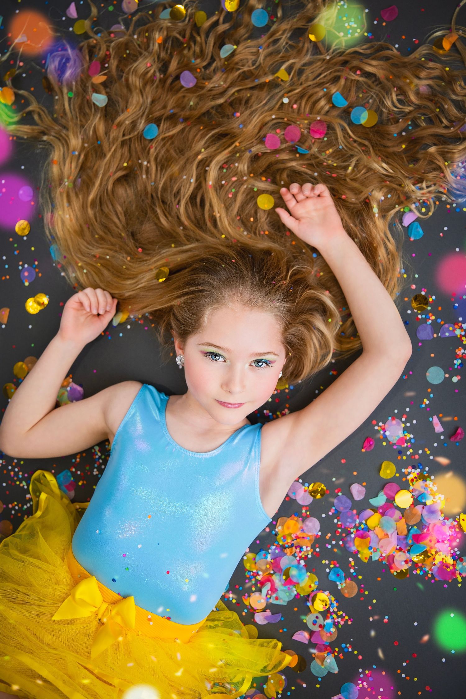 A young girl with long wavy hair lies on the floor in a blue top and yellow skirt with confetti around her.