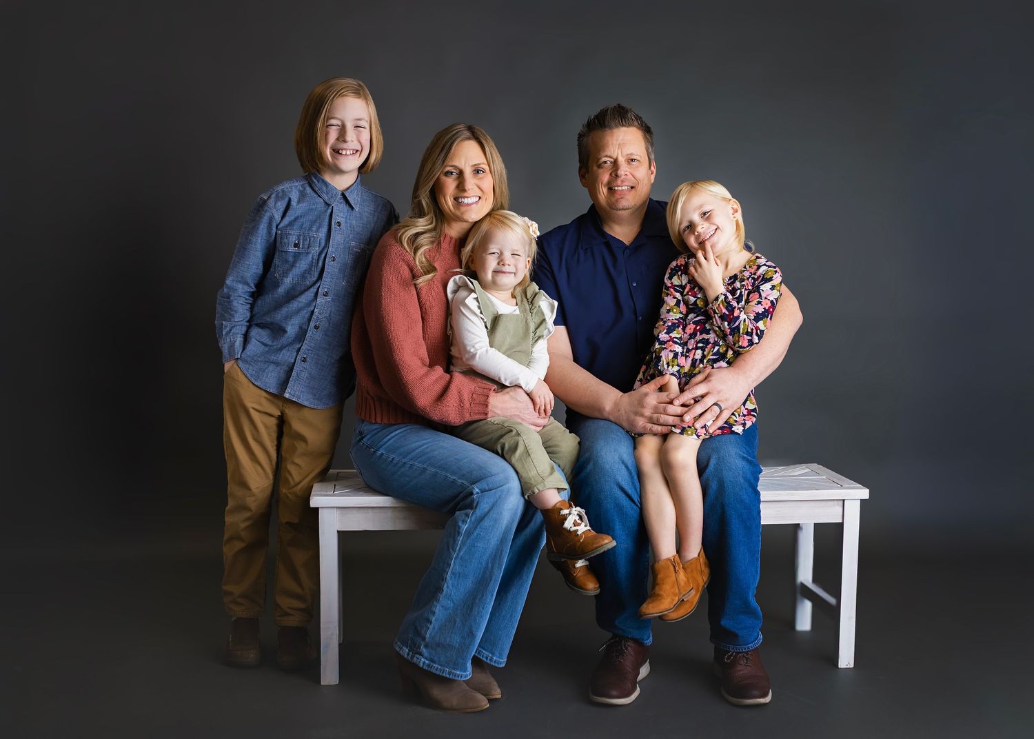 Family of five posing together on a white bench against a dark gray studio background.