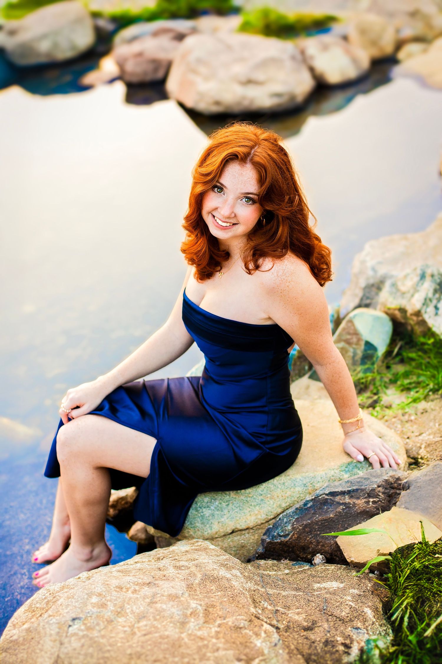 Smiling redhead woman in navy blue dress sitting on rocks beside a peaceful pond.