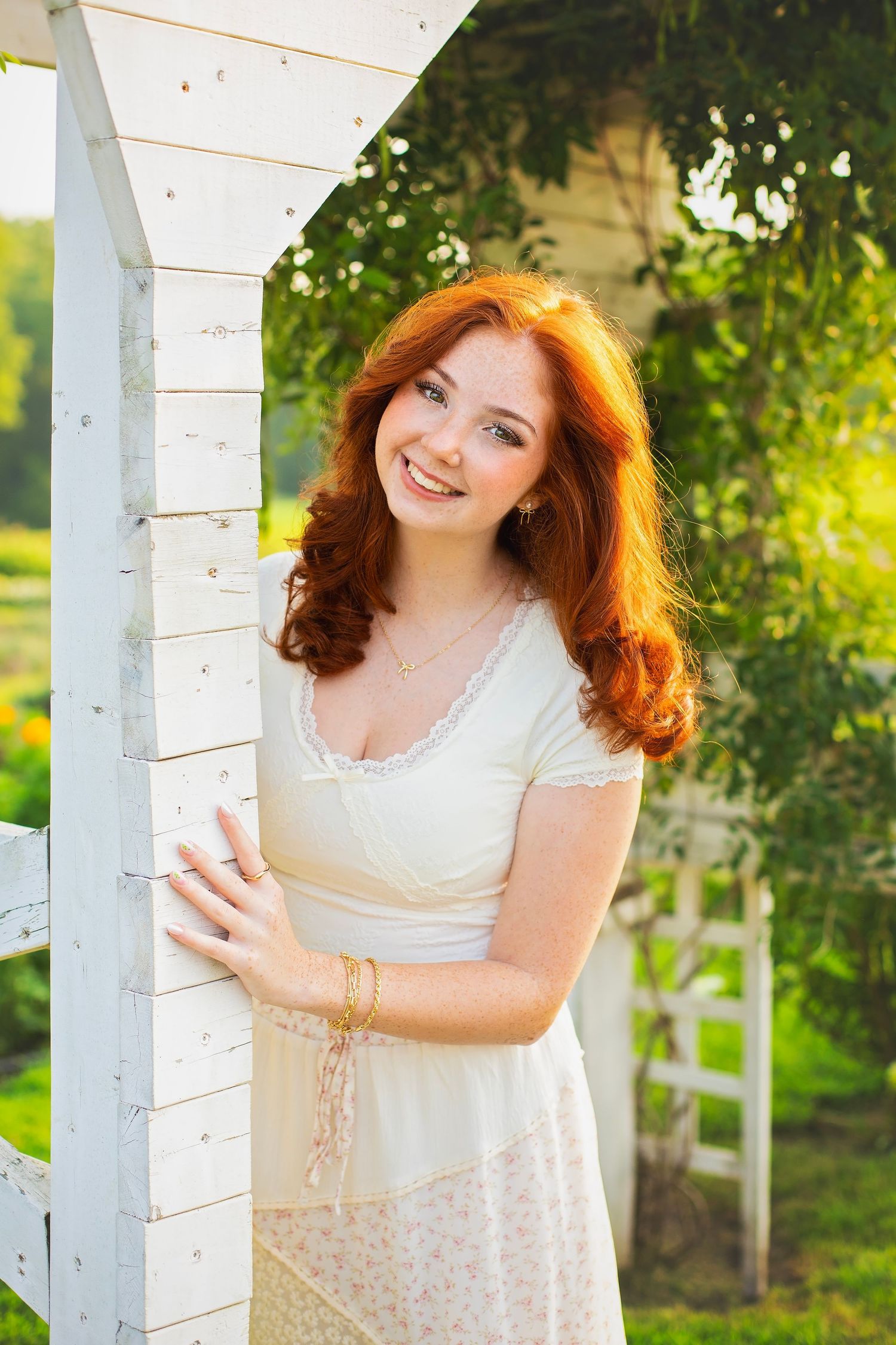 Smiling redhead teen in white dress leaning against a white garden arbor in sunlight.