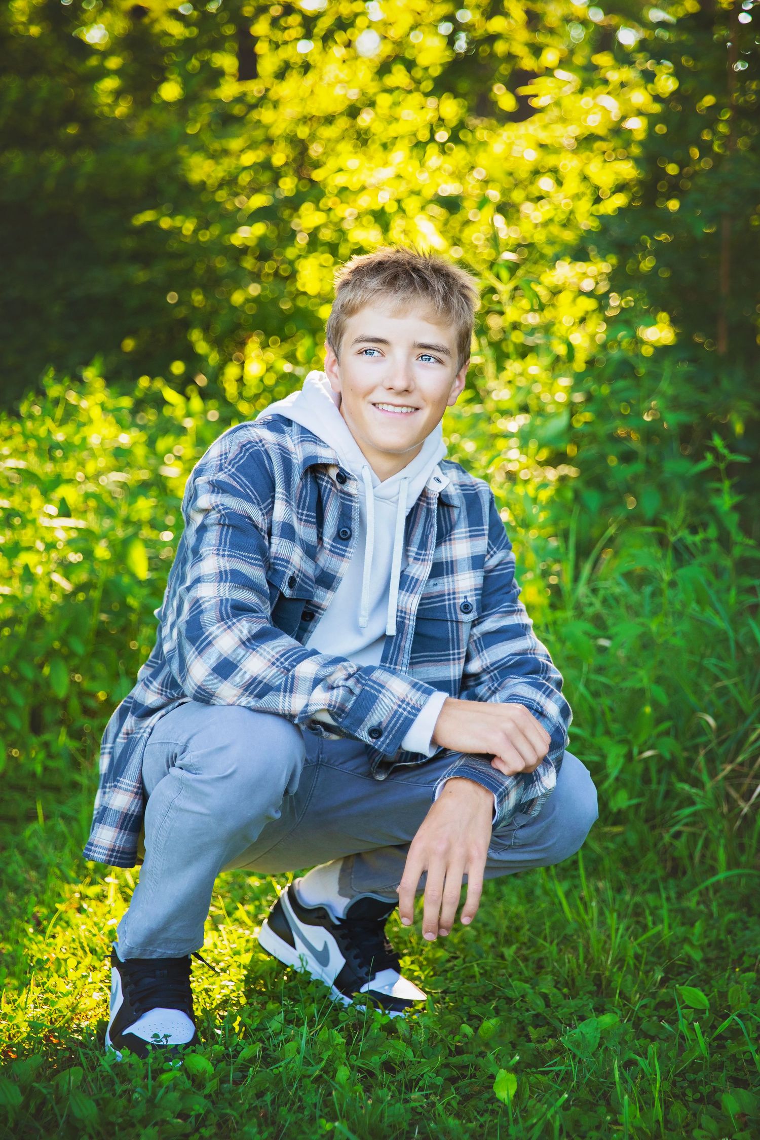 Smiling teenage boy crouching in green grass wearing a plaid flannel shirt over a hoodie.