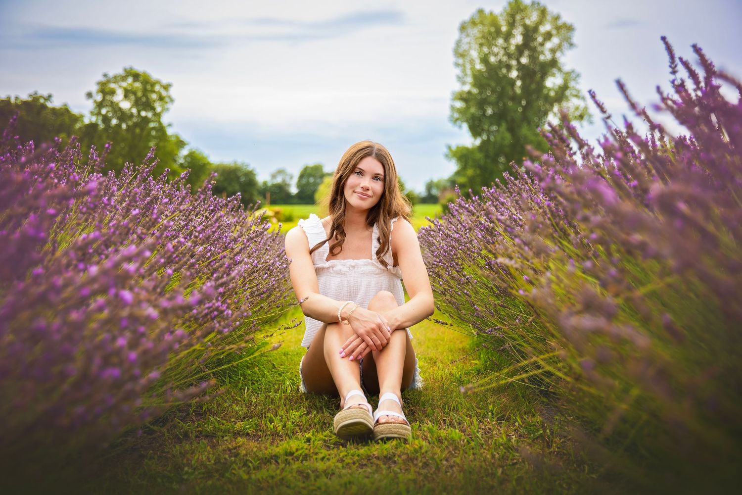 Teen girl in white dress sitting among purple lavender rows in a scenic field.