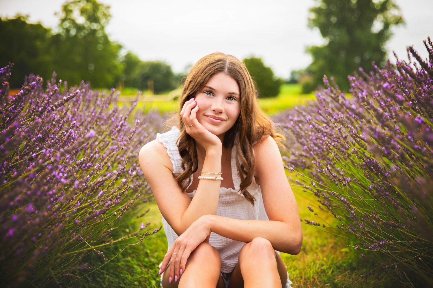 Teen girl smiling while seated among blooming purple lavender rows in a sunny field.