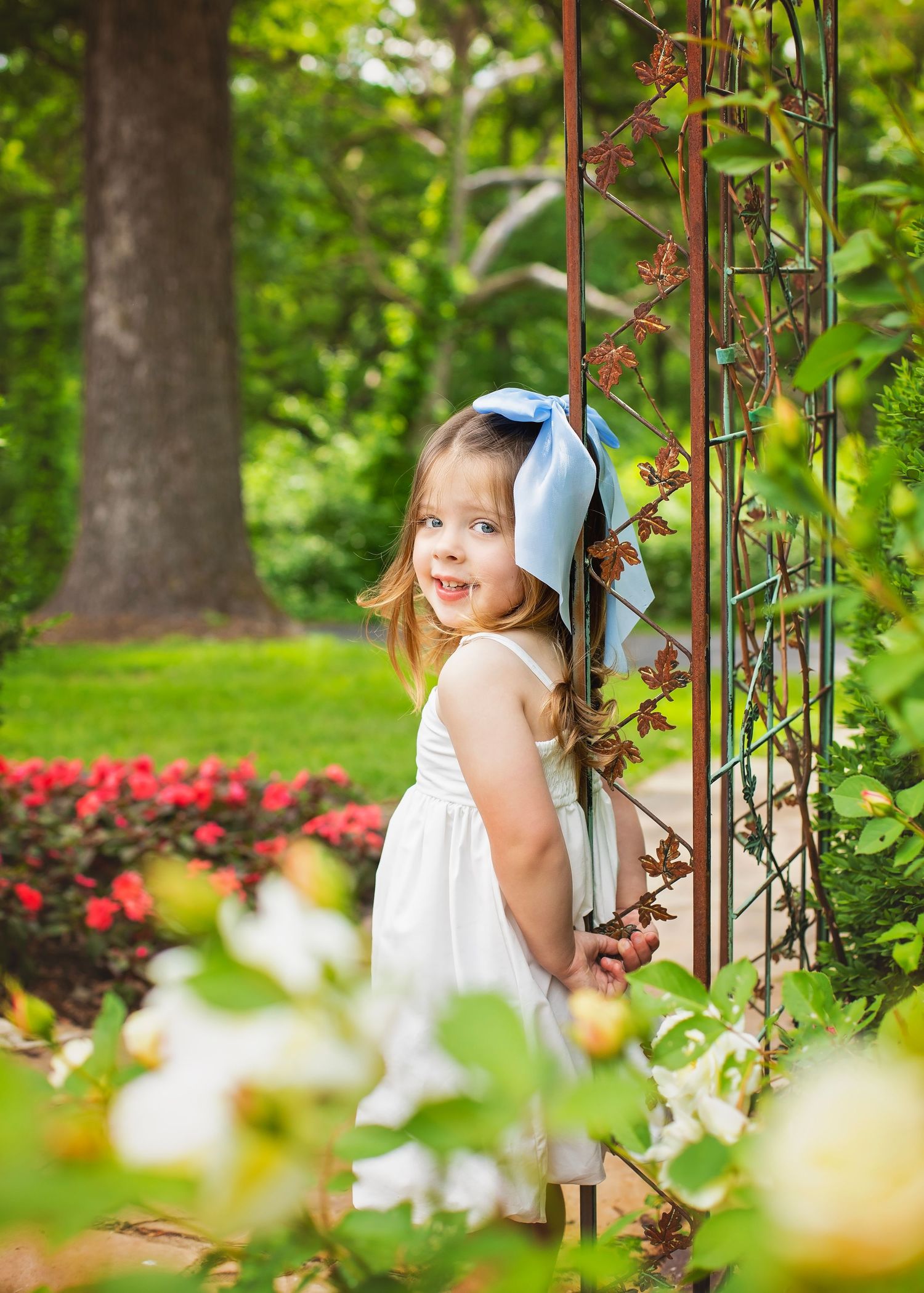Sweet little girl in white dress with blue bow standing by a garden trellis surrounded by colorful flowers.