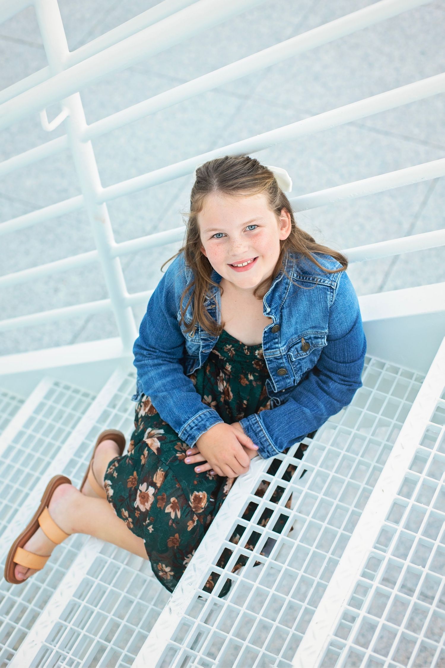 Young girl in a denim jacket and floral dress sitting on white outdoor stairs, smiling at the camera.