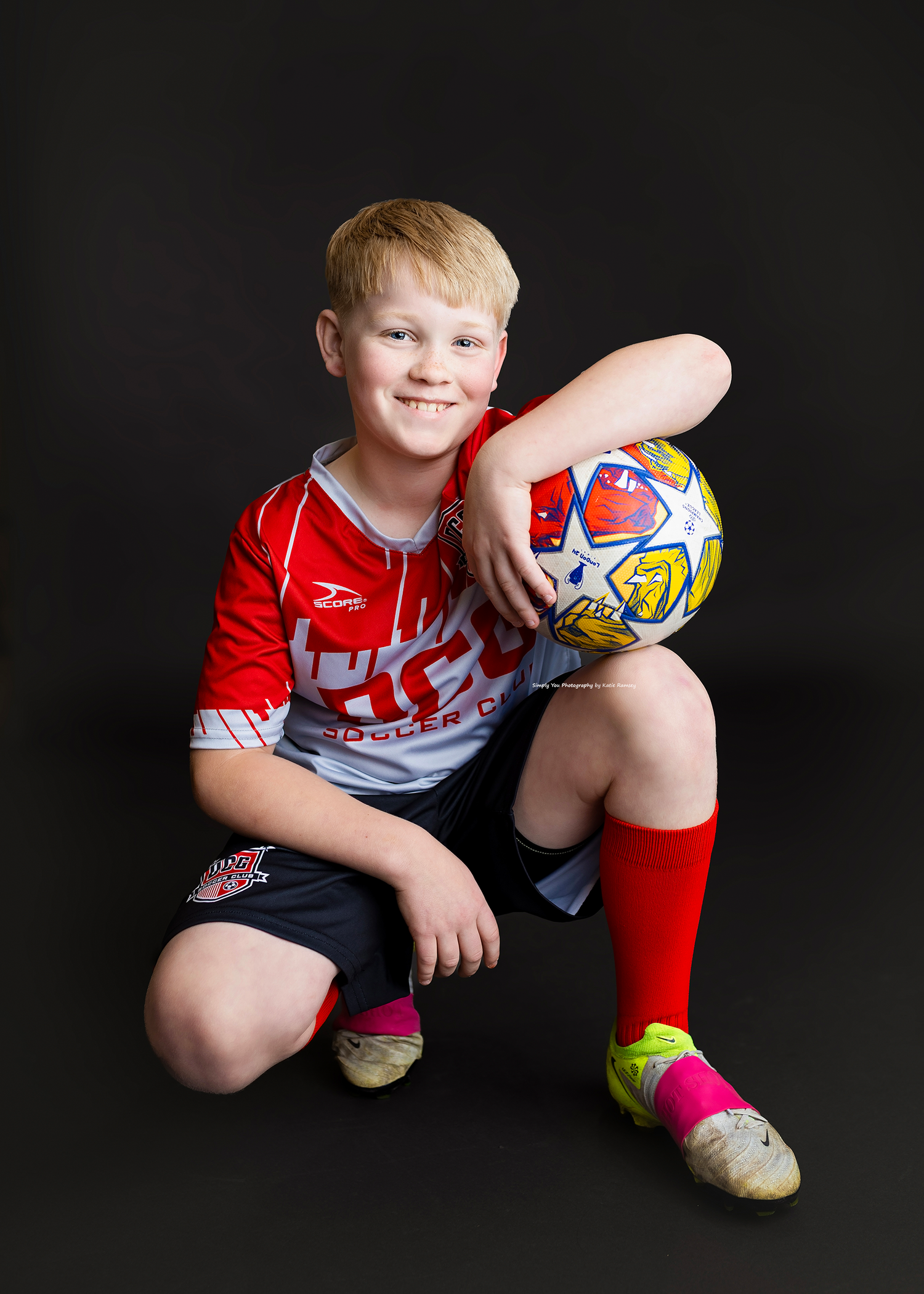 Young blonde boy in red soccer uniform poses with a soccer ball against a dark background.