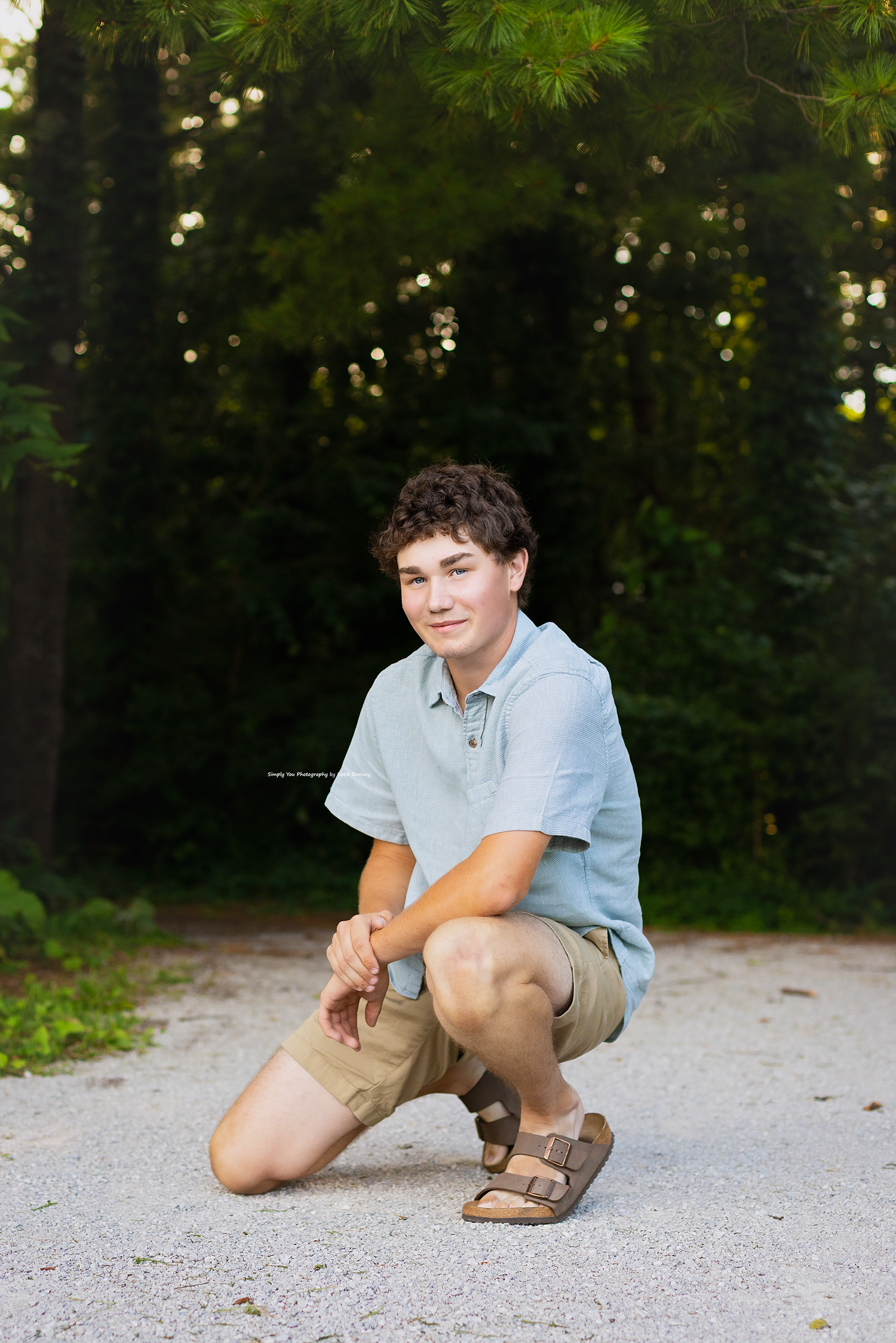 Teen boy in light blue polo and khaki shorts crouching on gravel path surrounded by lush green trees.