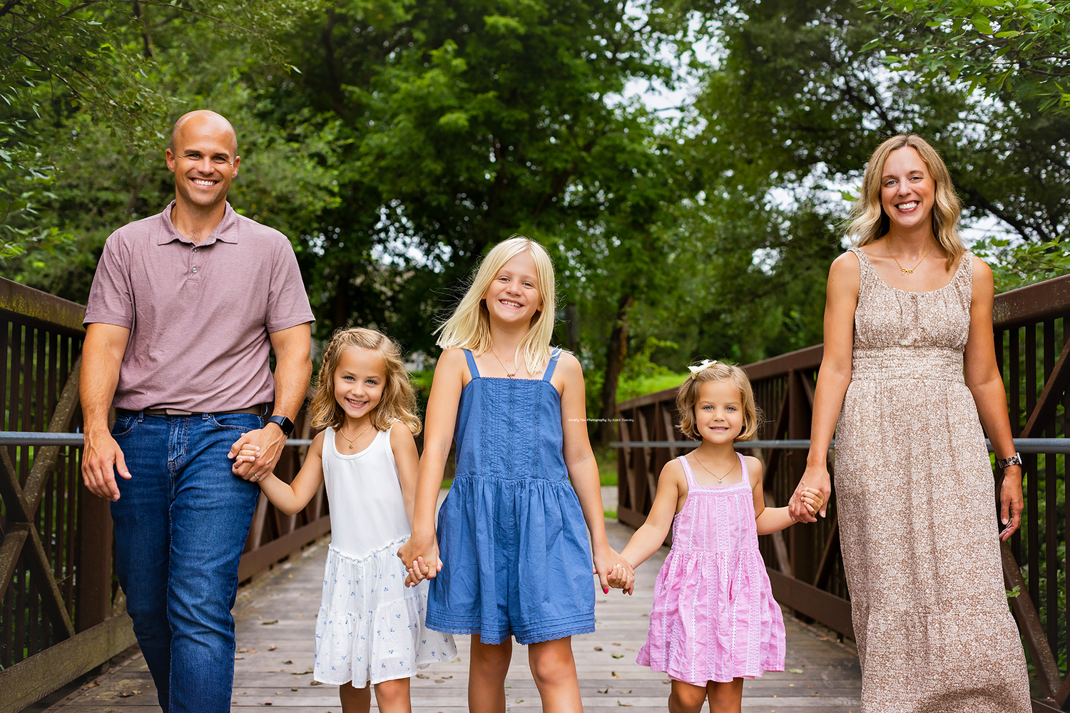 Happy family of five walking on a wooden bridge outdoors, parents holding hands with three young blonde daughters.