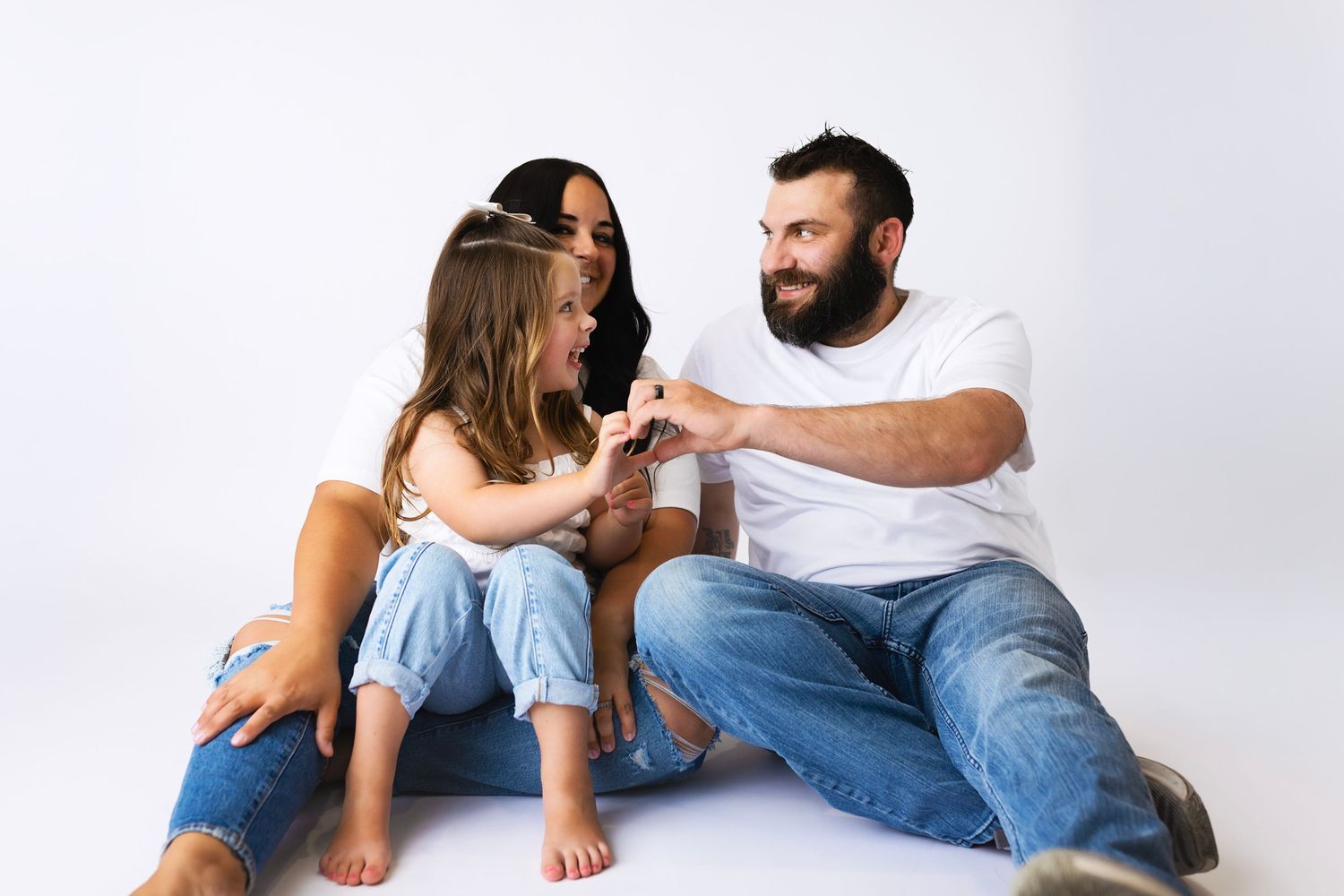 Family of three sitting on white floor, parents and young daughter making heart shapes with hands together.