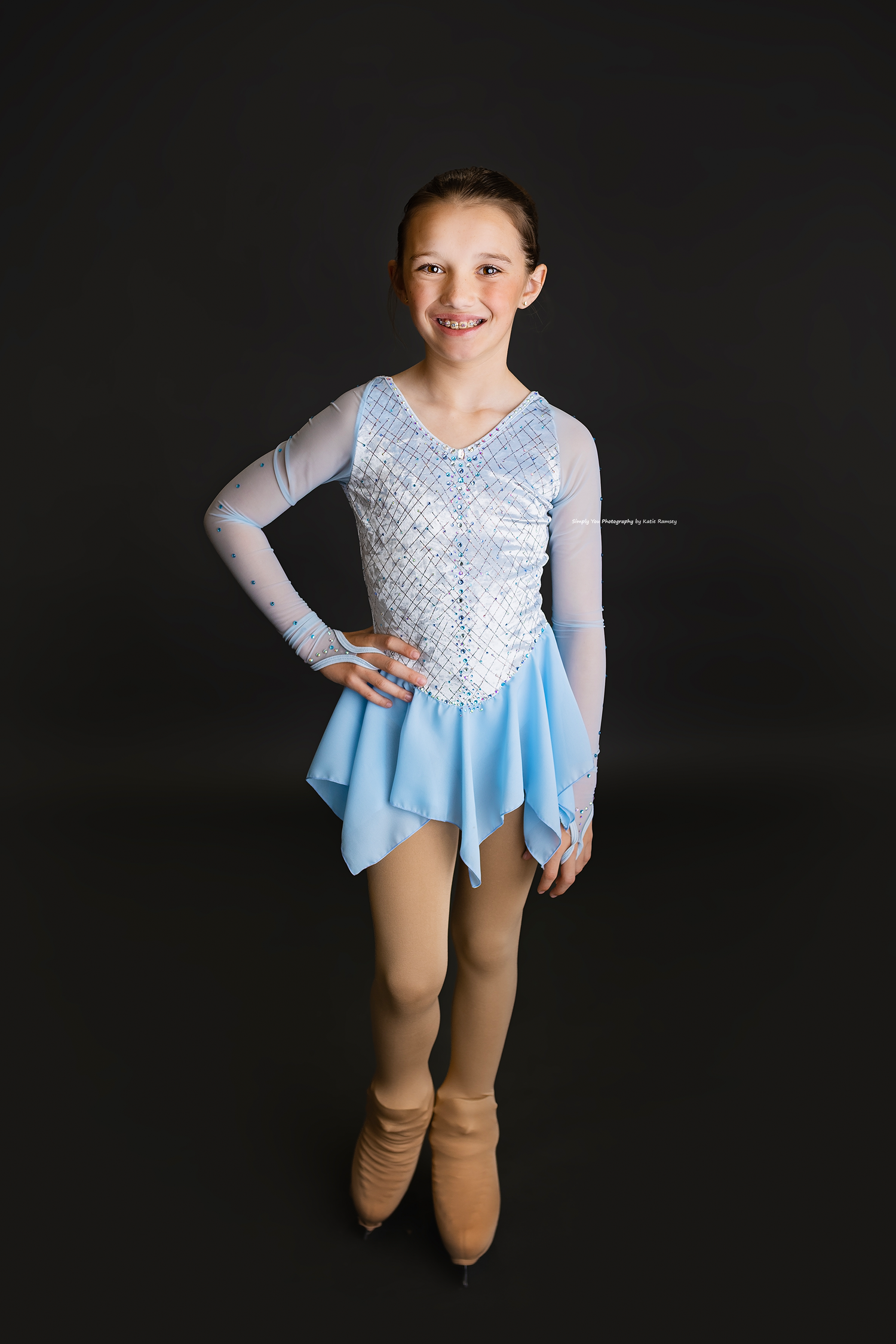 Young figure skater in light blue dress and white sequined bodice poses confidently on black background.