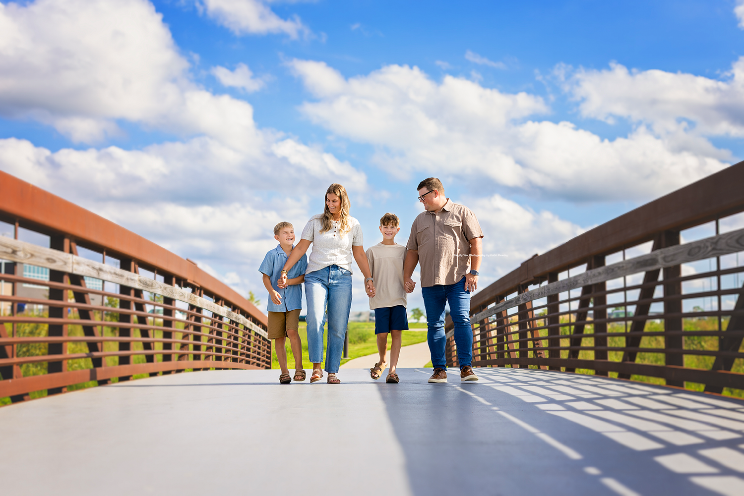 Family of four walking on a wooden bridge under a blue sky with white clouds on a sunny day.