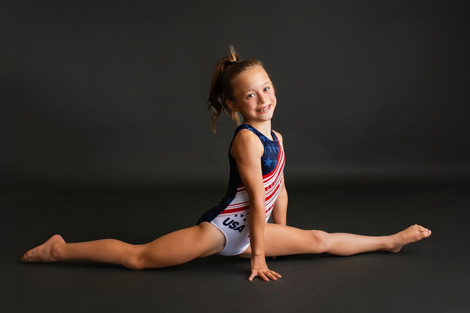 Young gymnast in USA leotard smiles while performing a full splits on the floor.