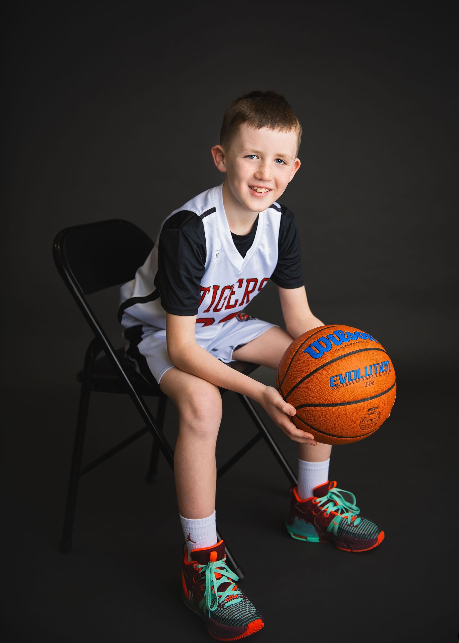 Smiling boy in Tigers basketball uniform sits on chair holding Wilson Evolution ball.