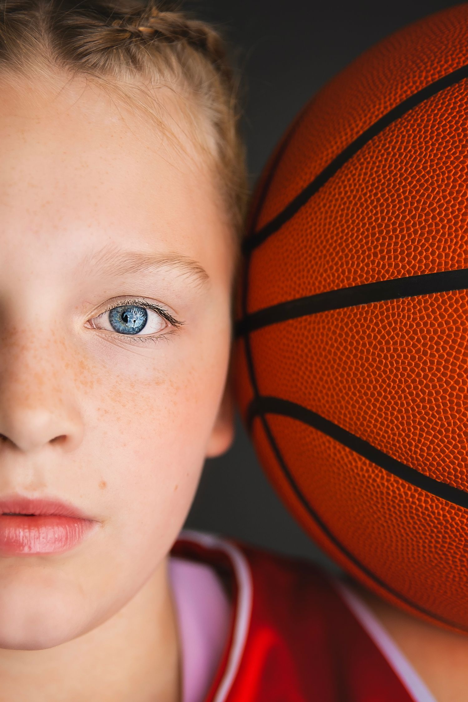 Close-up portrait of a young girl with blue eyes and freckles holding a basketball beside her face.