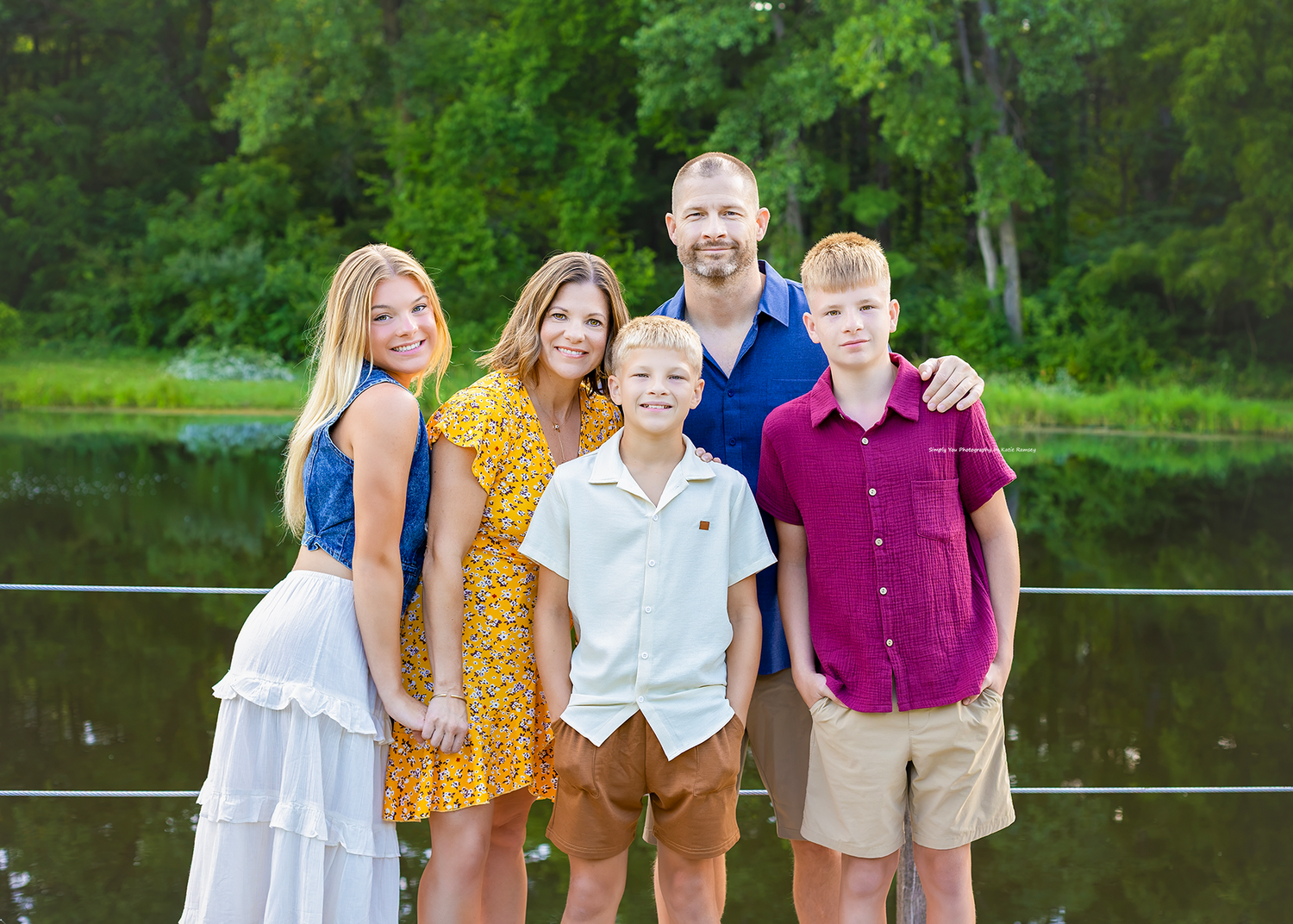 Family of five posing outdoors near a pond surrounded by lush green trees on a sunny day.